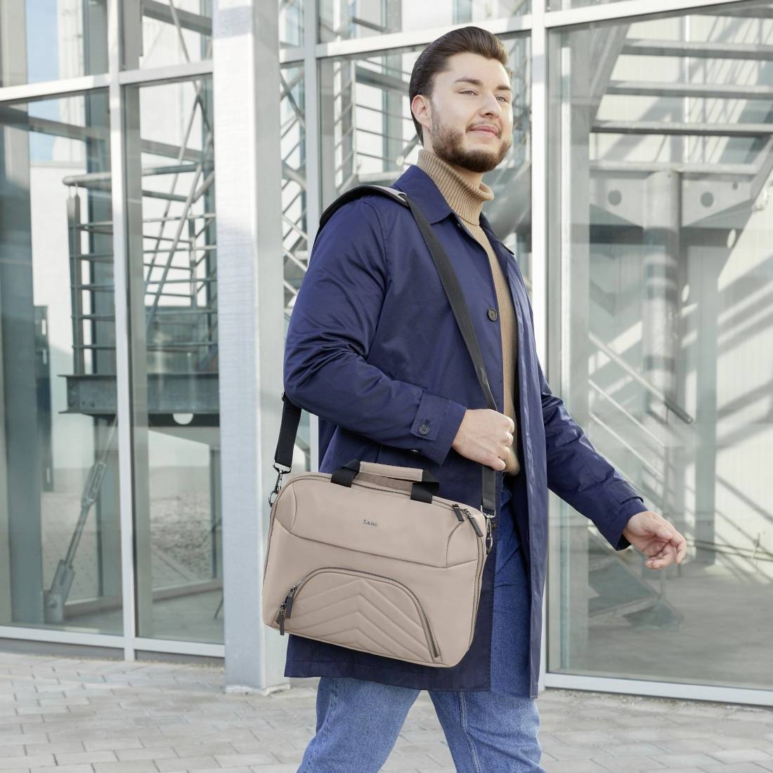 Een man draagt een blauwe jas en houdt een lichte schoudertas vast. Hij staat voor een modern, glazen gebouw.