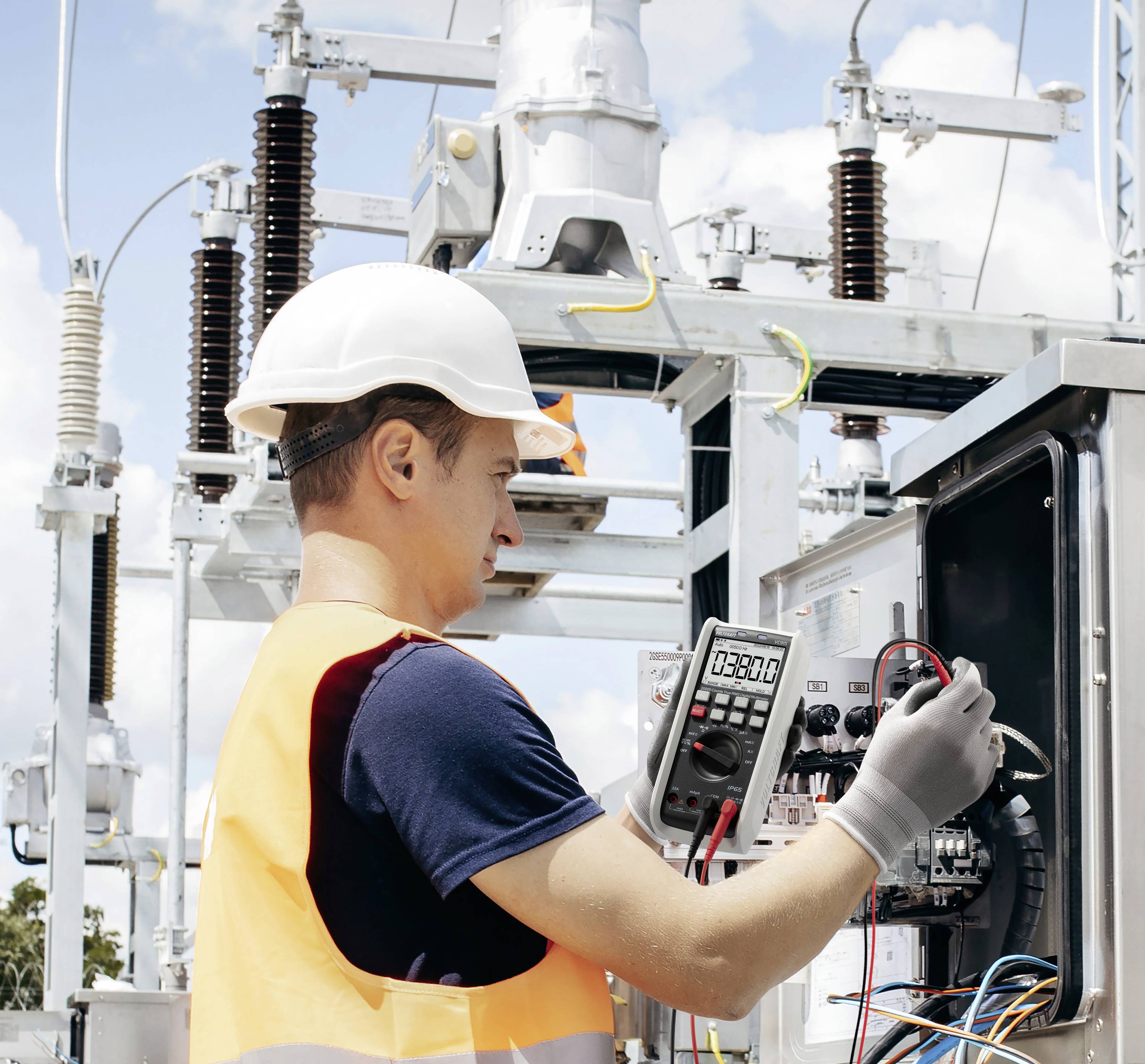 'Technicus met een veiligheidshelm en veiligheidshesje gebruikt een multimeter om elektrische apparatuur te controleren bij een openlucht-hoogspanningsstation.'