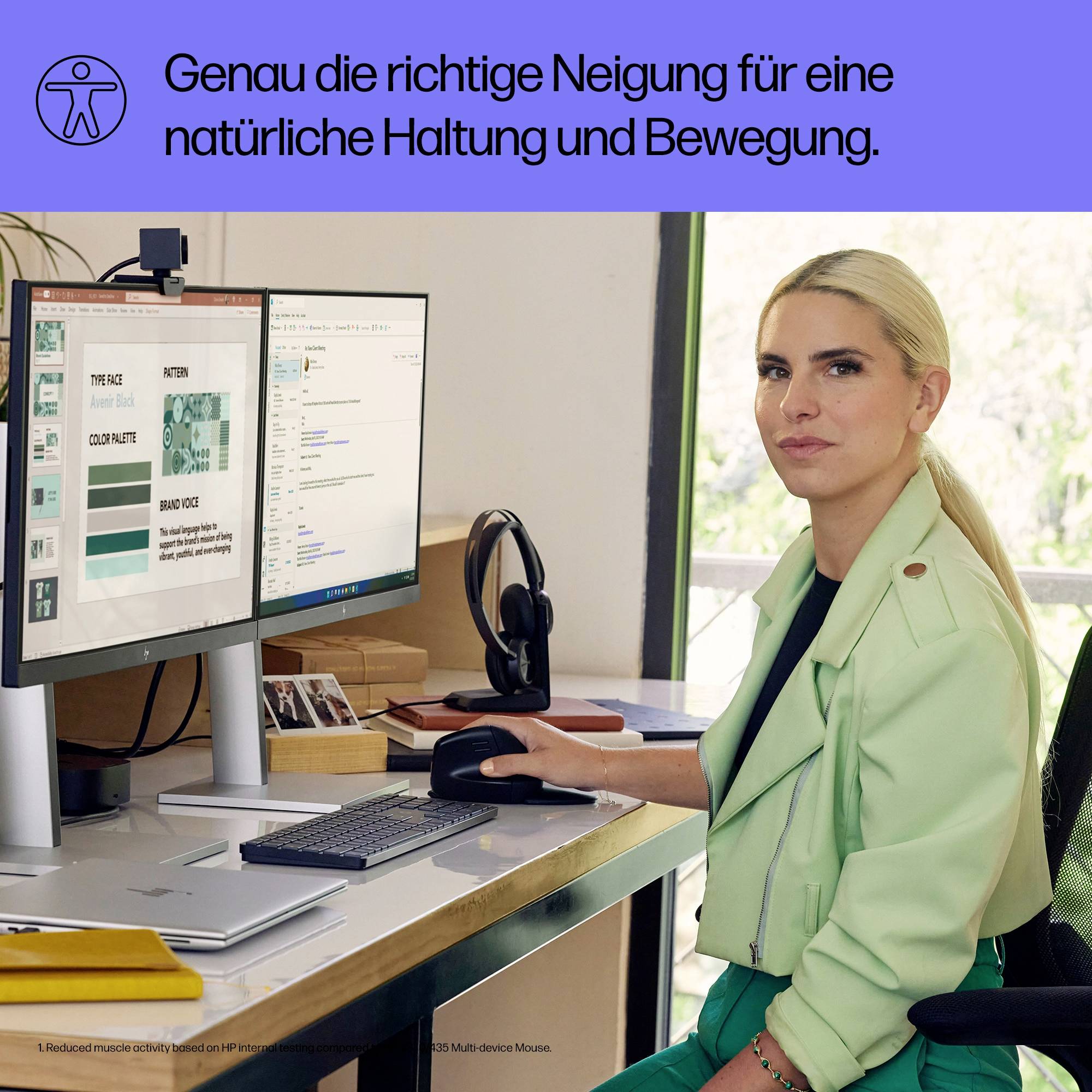 Vrouw in een groene jas zit aan een bureau met twee schermen, een kopje koffie, toetsenbord en muis. Op de achtergrond planten.