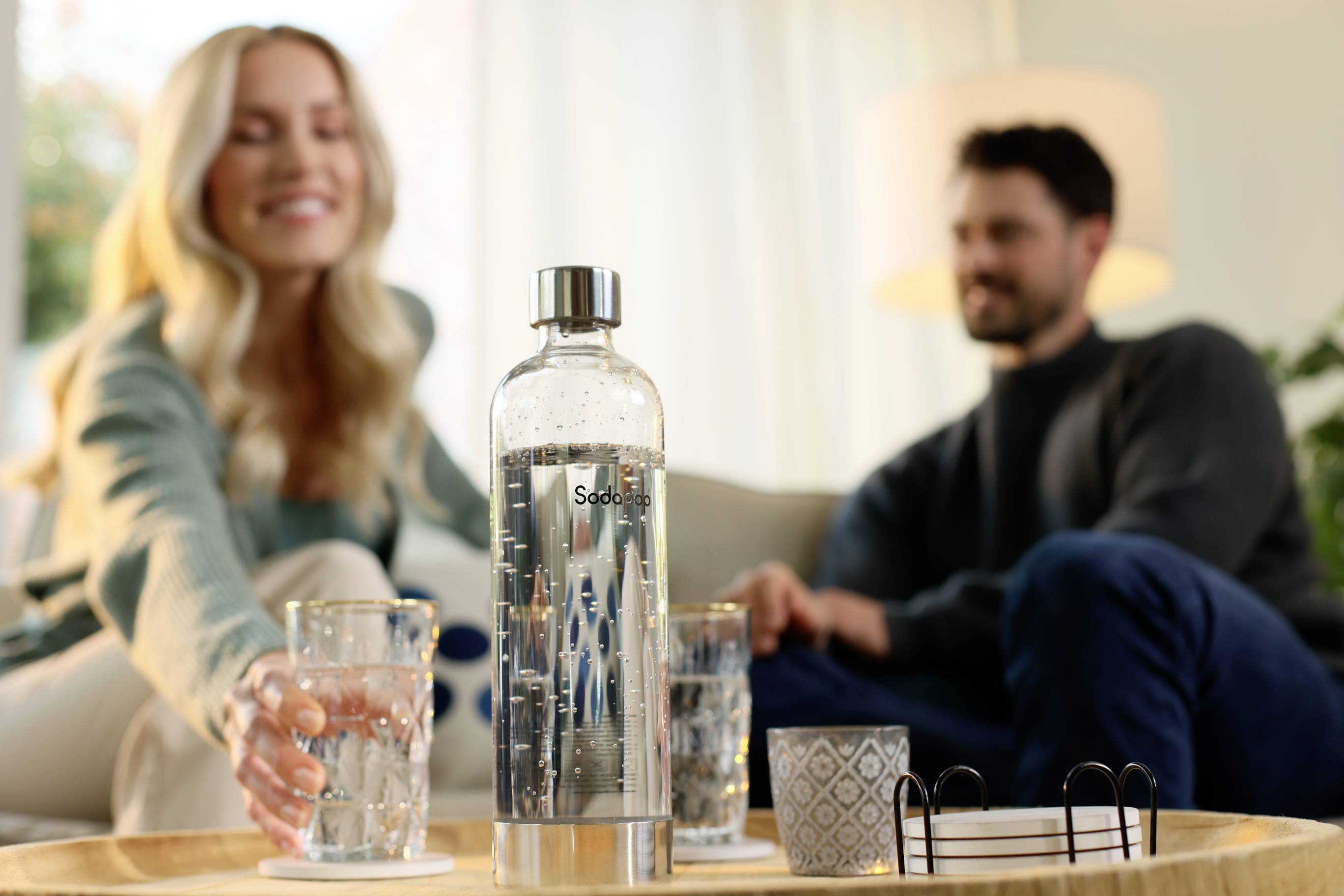 Een vrouw reikt een man een glas water in een woonkamer. Op de voorgrond staat een met water gevulde Sodastream-fles.
