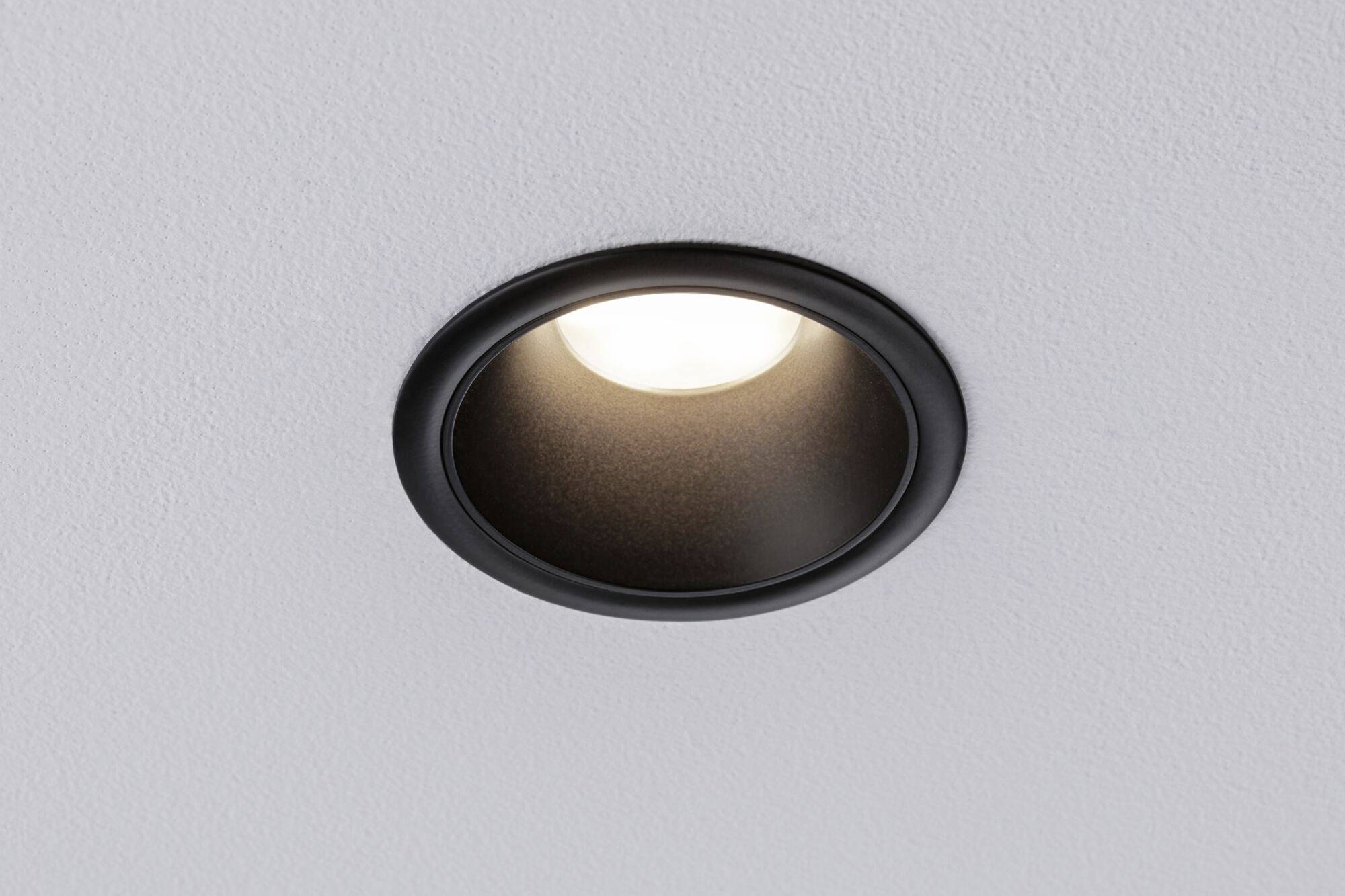 Een zwarte, ronde inbouwspot in een witte plafond, die is ingeschakeld en warm licht uitstraalt.