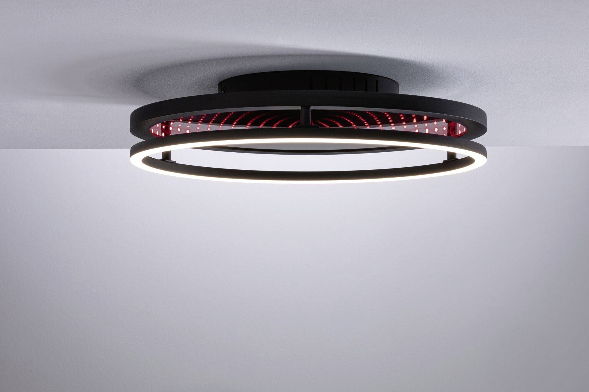 Een moderne plafondlamp met twee cirkelvormige verlichtingsringen die warm licht uitstralen. Zwarte structuur met rood binnengebied.