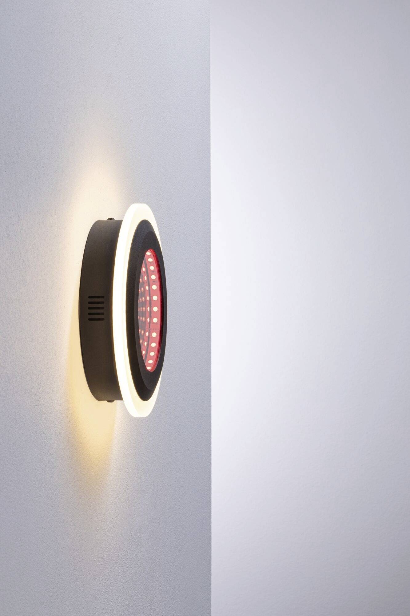 Wandlamp met zwarte behuizing, een rood LED-lichtcentrum en een witte verlichtingsring, gemonteerd op een witte muur.