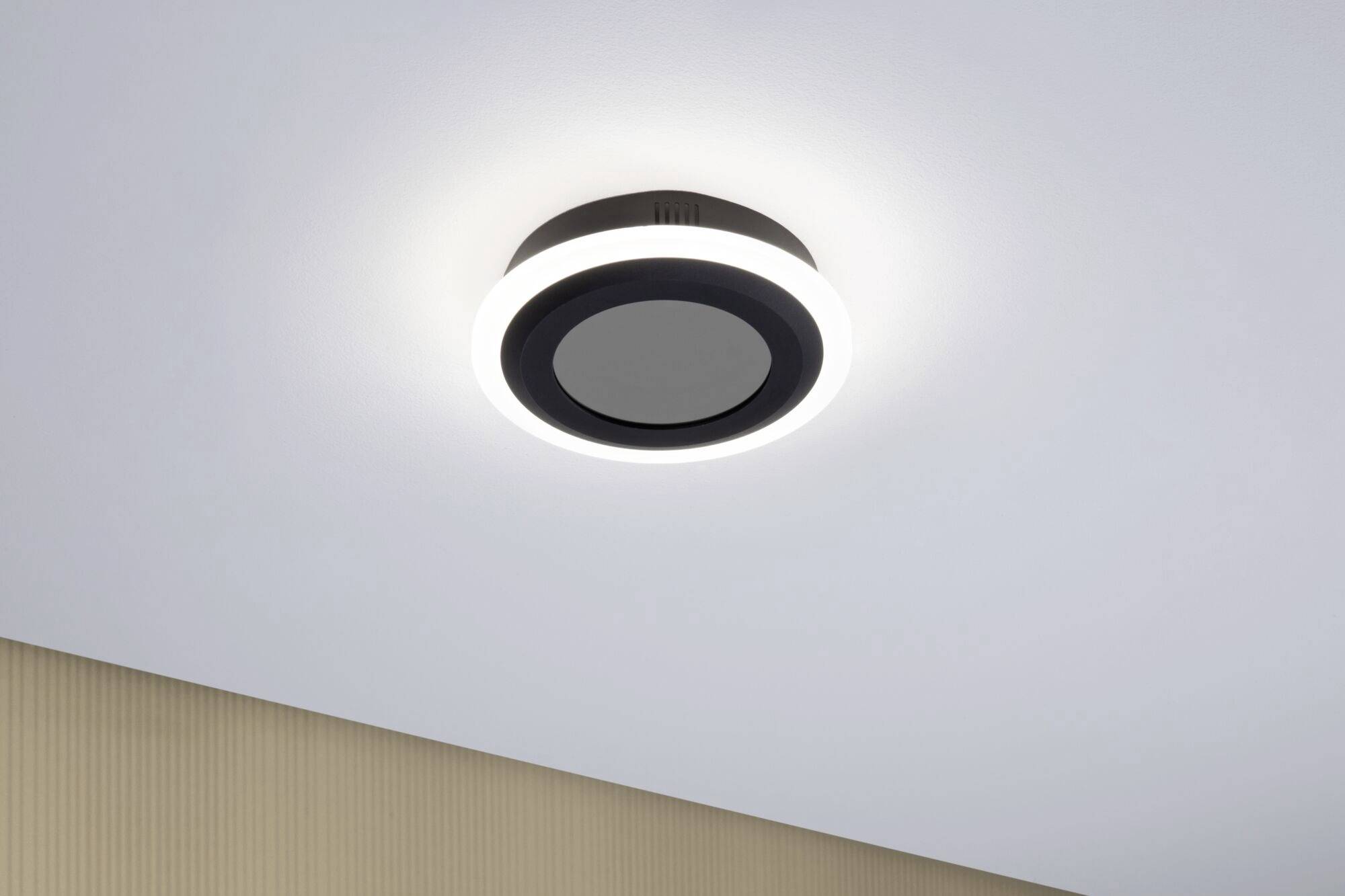 Zwarte, ronde plafondlamp met verlichte LED-ring, gemonteerd aan een witte plafond boven een beige muur. Modern design.