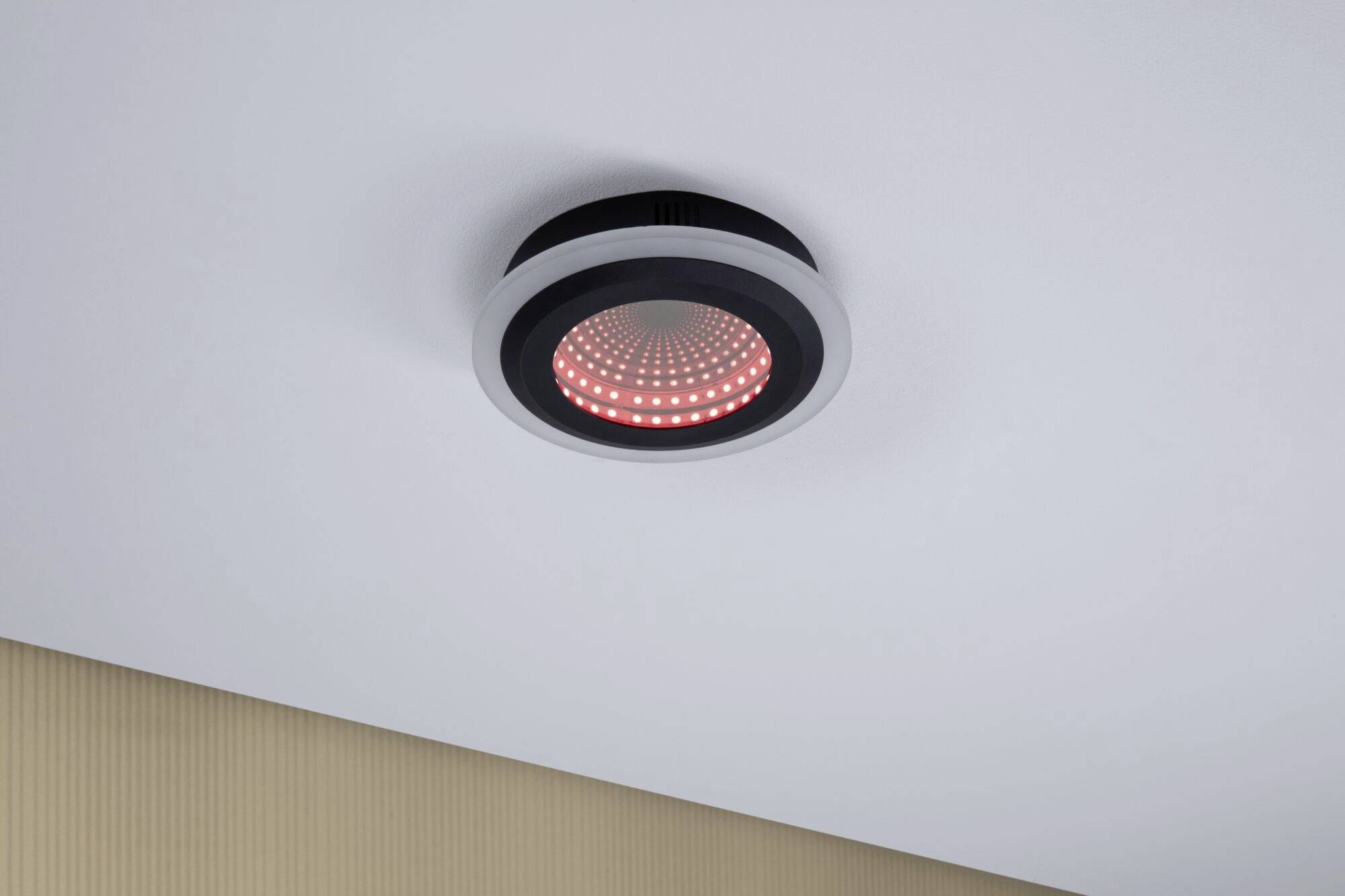 Een zwart-grijze plafondlamp met rood led-licht is gemonteerd aan een witte plafond. Hij verlicht een moderne ruimte.