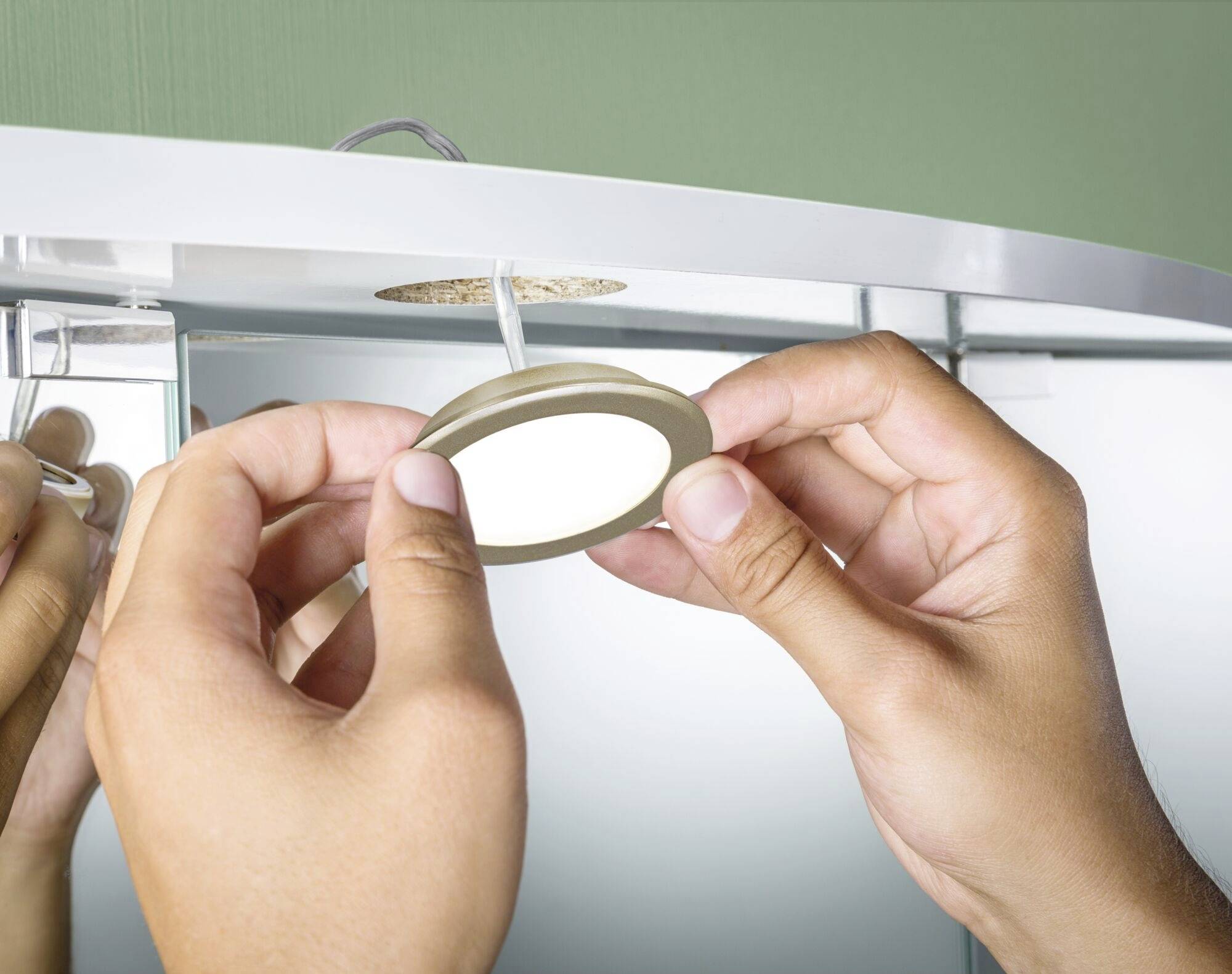 Een persoon installeert een rond, plat LED-licht in een metalen oppervlak, waarschijnlijk onder een keukenkastje.