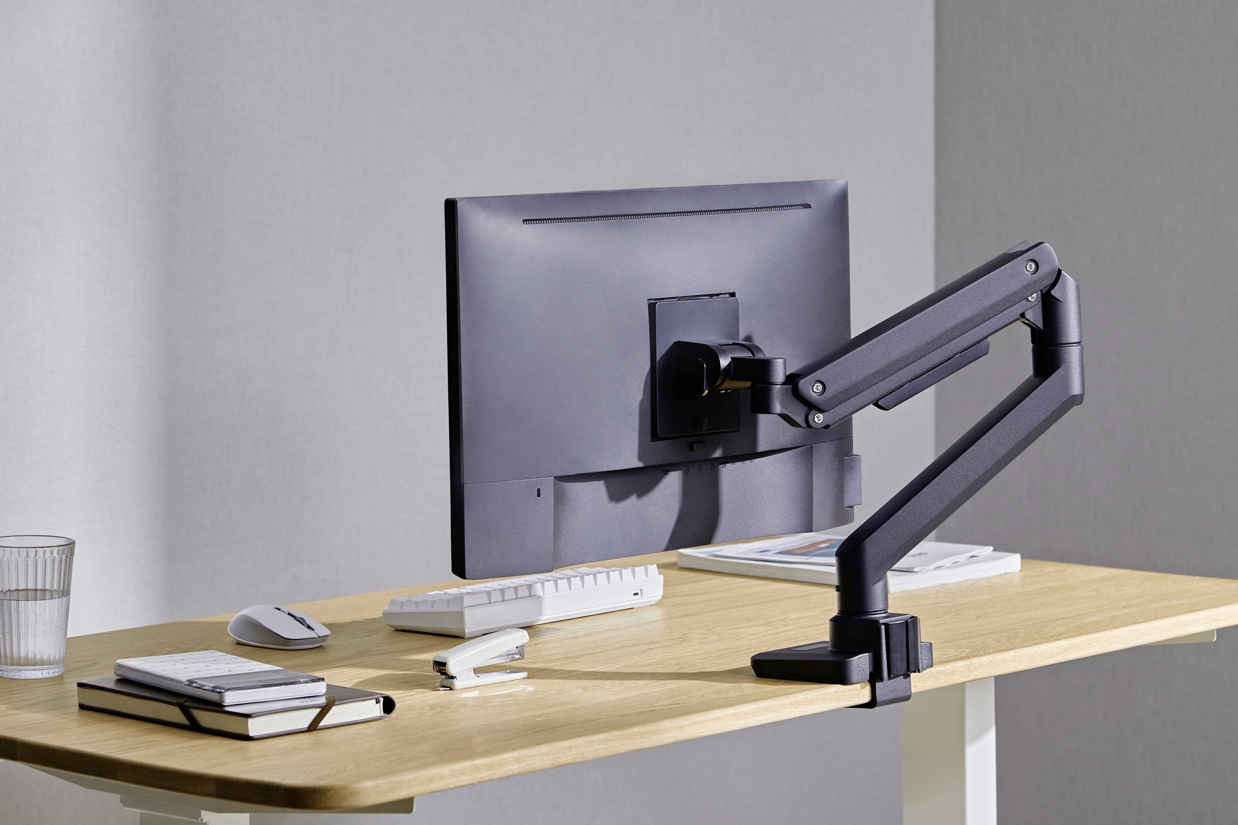 Een minimalistische werkplek met een monitor op een verstelbare arm, naast een toetsenbord, muis, stapel notitieboeken en een glas water.