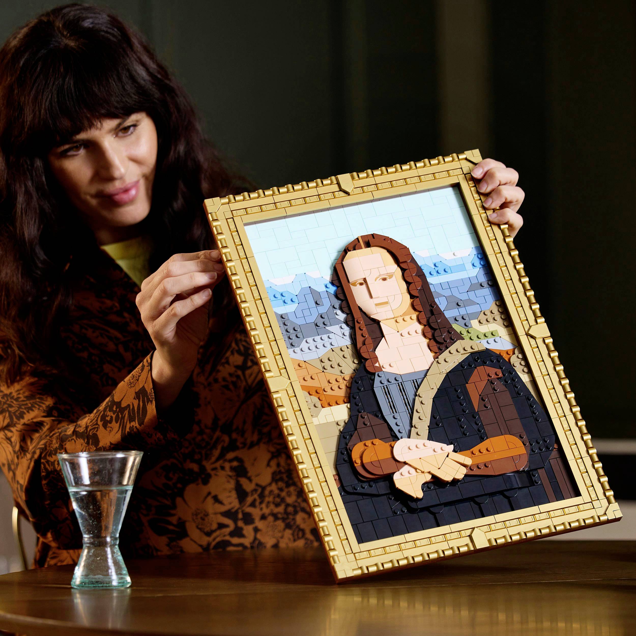 Een persoon houdt een mozaïekafbeelding van de Mona Lisa gemaakt van kleurrijke Lego-blokjes vast en bekijkt deze. Een glas water staat op tafel.