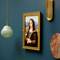 Een Legoschilderij van de Mona Lisa hangt aan een turquoise muur, geflankeerd door een groene lamp links en gouden decoratieve elementen rechts.