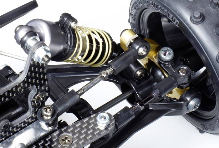 Close-up van een modelauto-vering met een spiraalvormige schokdemper, metalen armen en een deel van een grote band op de achtergrond.