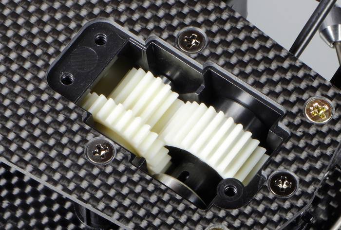 Twee witte tandwielen in een zwarte behuizing, gemonteerd op een koolstofvezelversterkte plaat, zichtbaar van bovenaf.