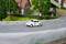 Een kleine witte speelgoedauto met nummer 53 rijdt over een modelstraatbaan langs gemodelleerde huizen en bomen.