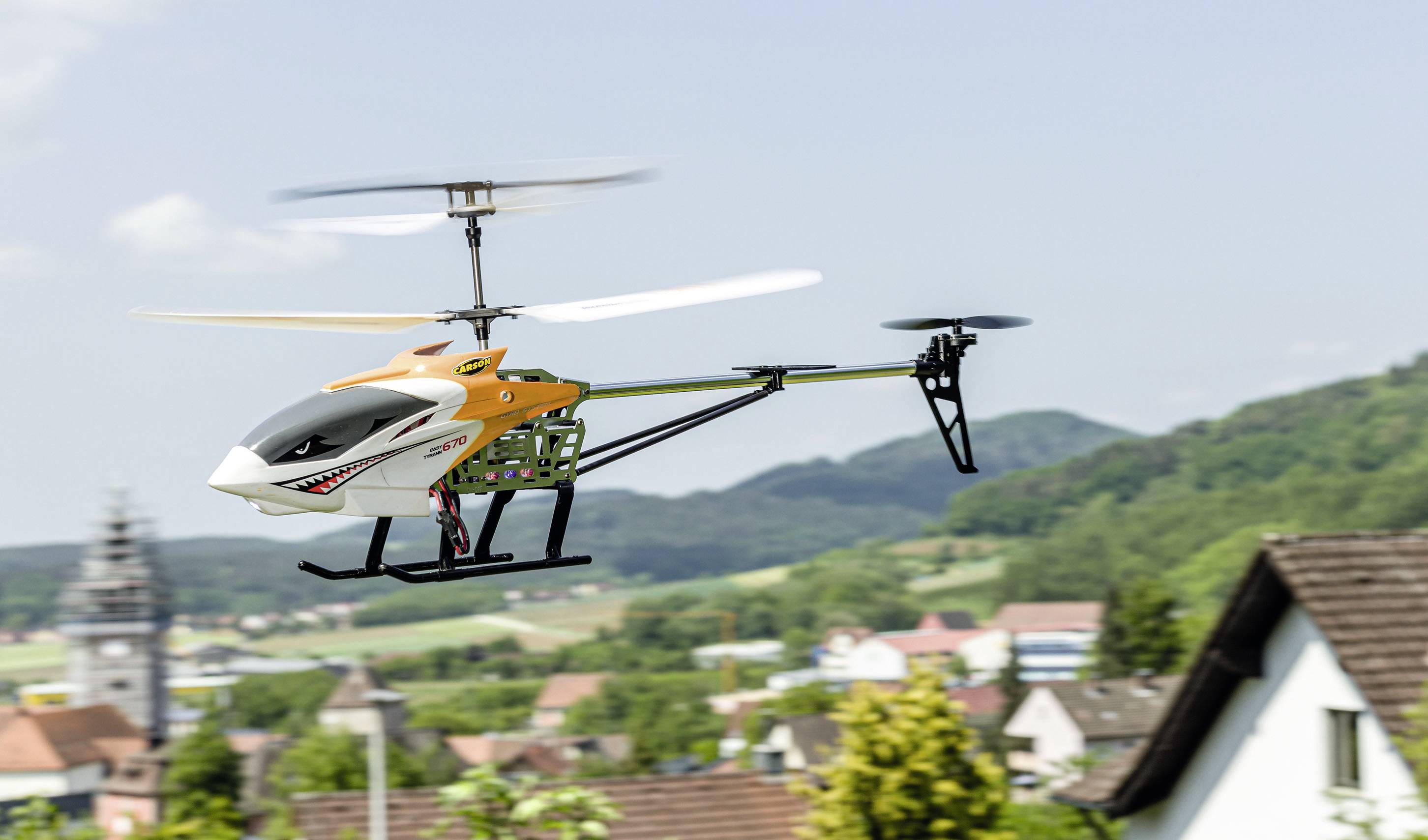 Een afstandsbestuurde helikopter vliegt over een landelijk stadslandschap met huizen en heuvels op de achtergrond.