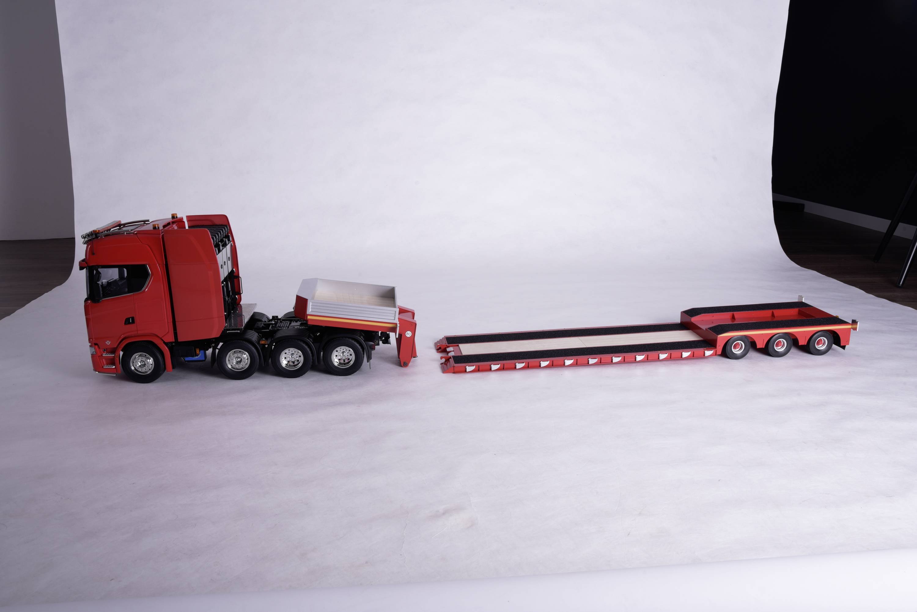 Een rood speelgoedmodel van een vrachtwagen met een lange dieplader, neergezet op een witte achtergrond.