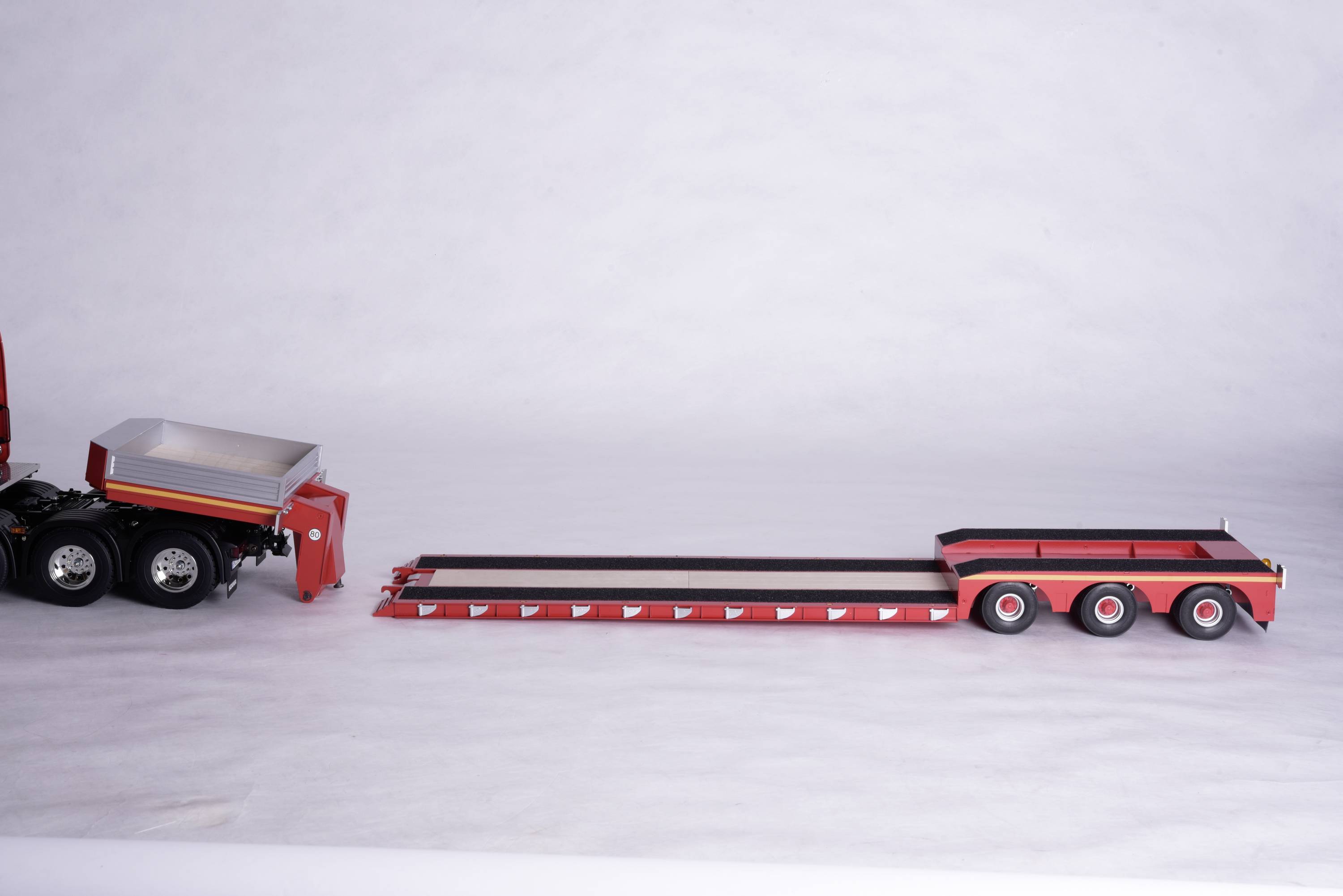 Een rood, drieassig dieplader-aanhangermodel is afgebeeld op een lichte ondergrond. Slechts een deel van een vrachtwagen is zichtbaar.
