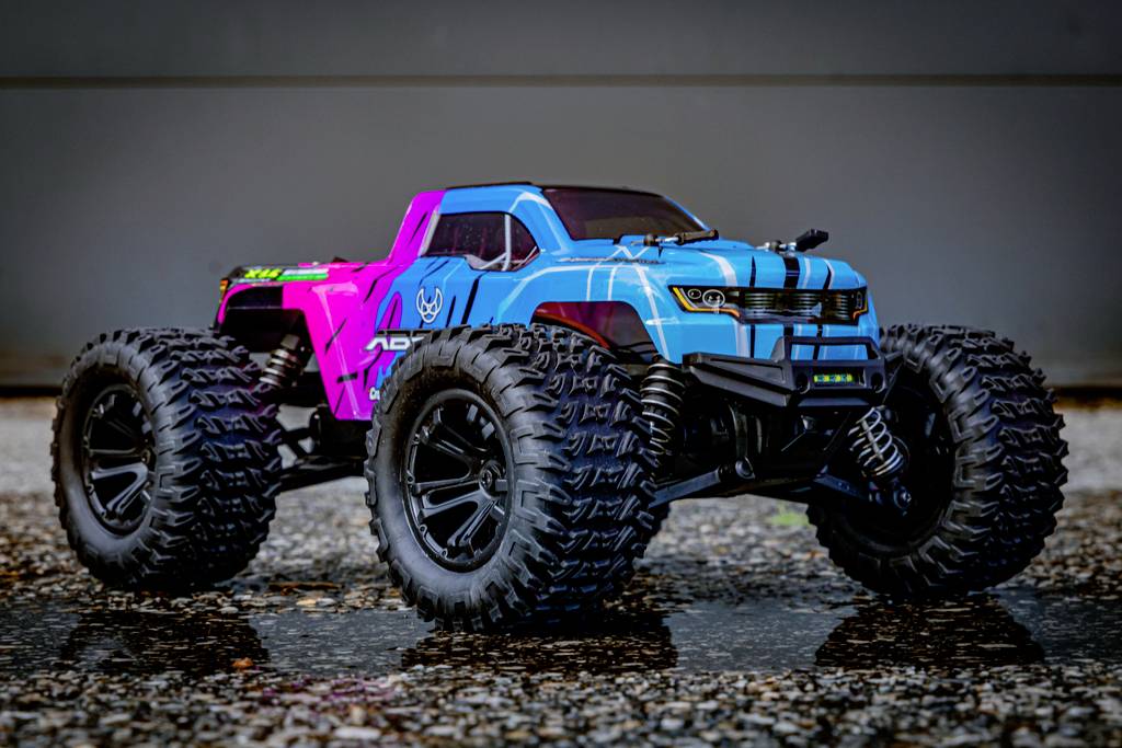 Een afstandsbediende auto staat op een natte asfaltweg. Hij is blauw en roze met grote, grof geprofilereerde banden, geschikt voor terreinrijden.
