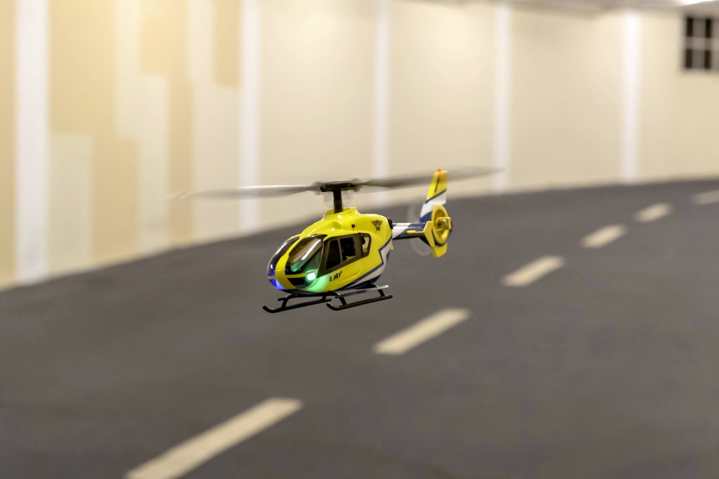 Een gele modelhelikopter vliegt in een binnenruimte boven een donker oppervlak met witte lijnen die een weg voorstellen.