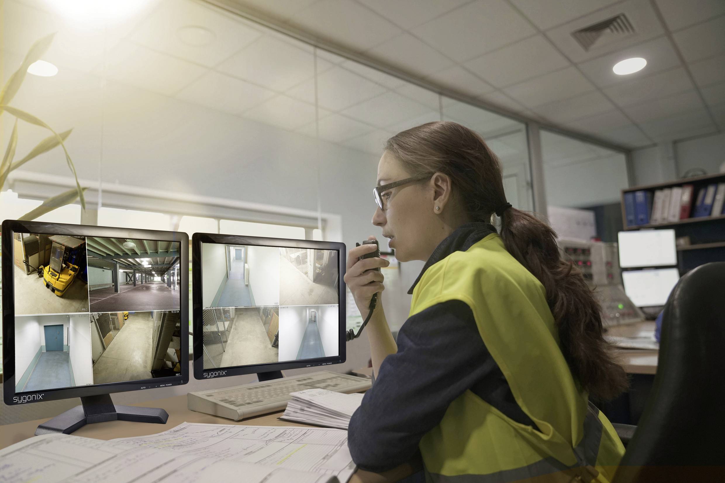 Een vrouw met een gele veiligheidsvest zit aan een bureau en houdt toezicht op videocamera-beelden op twee monitors in een controlekamer.