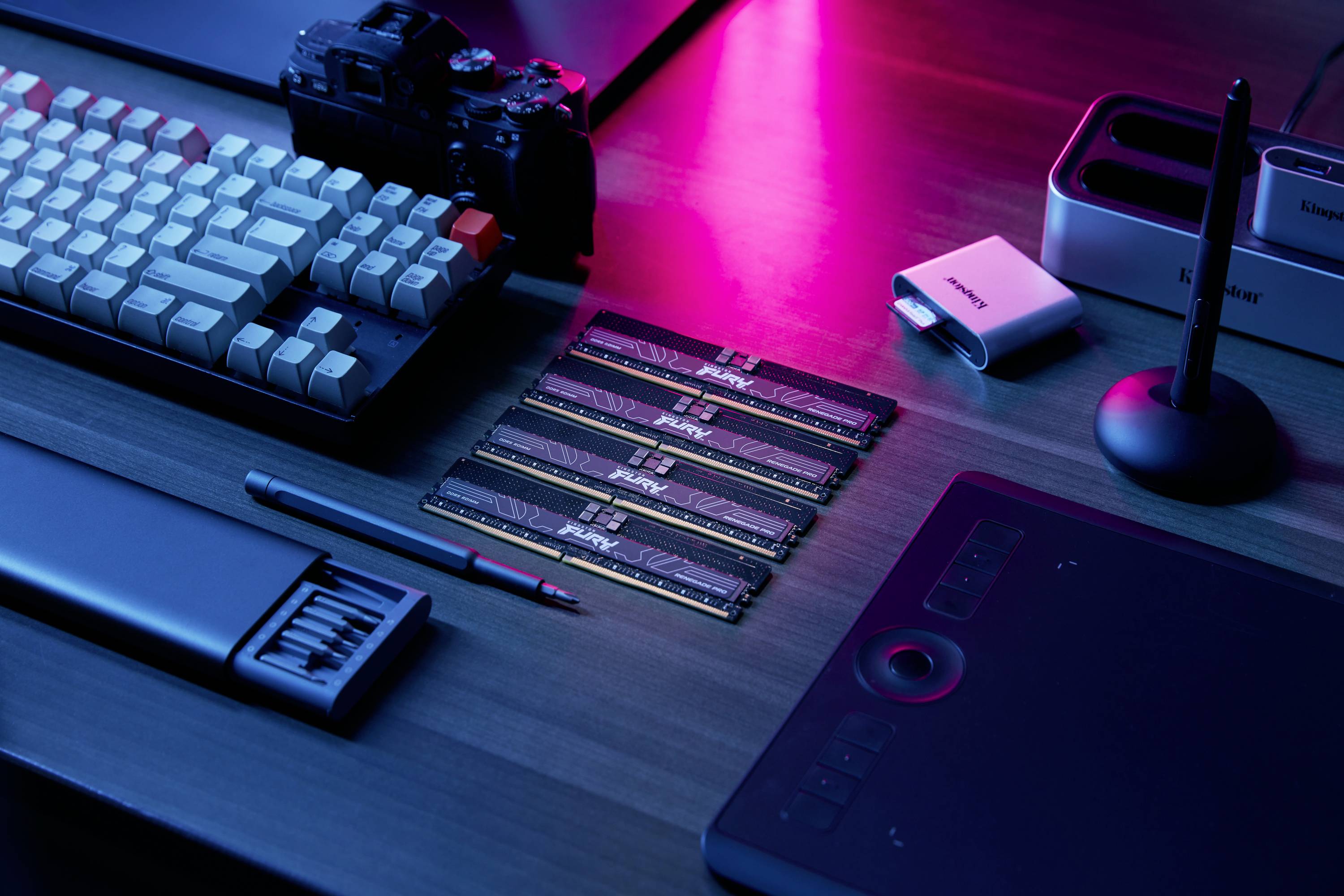 'Computerarbeidsplaats met toetsenbord, grafisch tablet, camera en RAM-modules op een tafel; roze en blauw verlicht.'