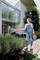 Een vrouw in tuinbroek besproeit planten met een tuinslang voor een modern huis met glazen wand op een houten terras.