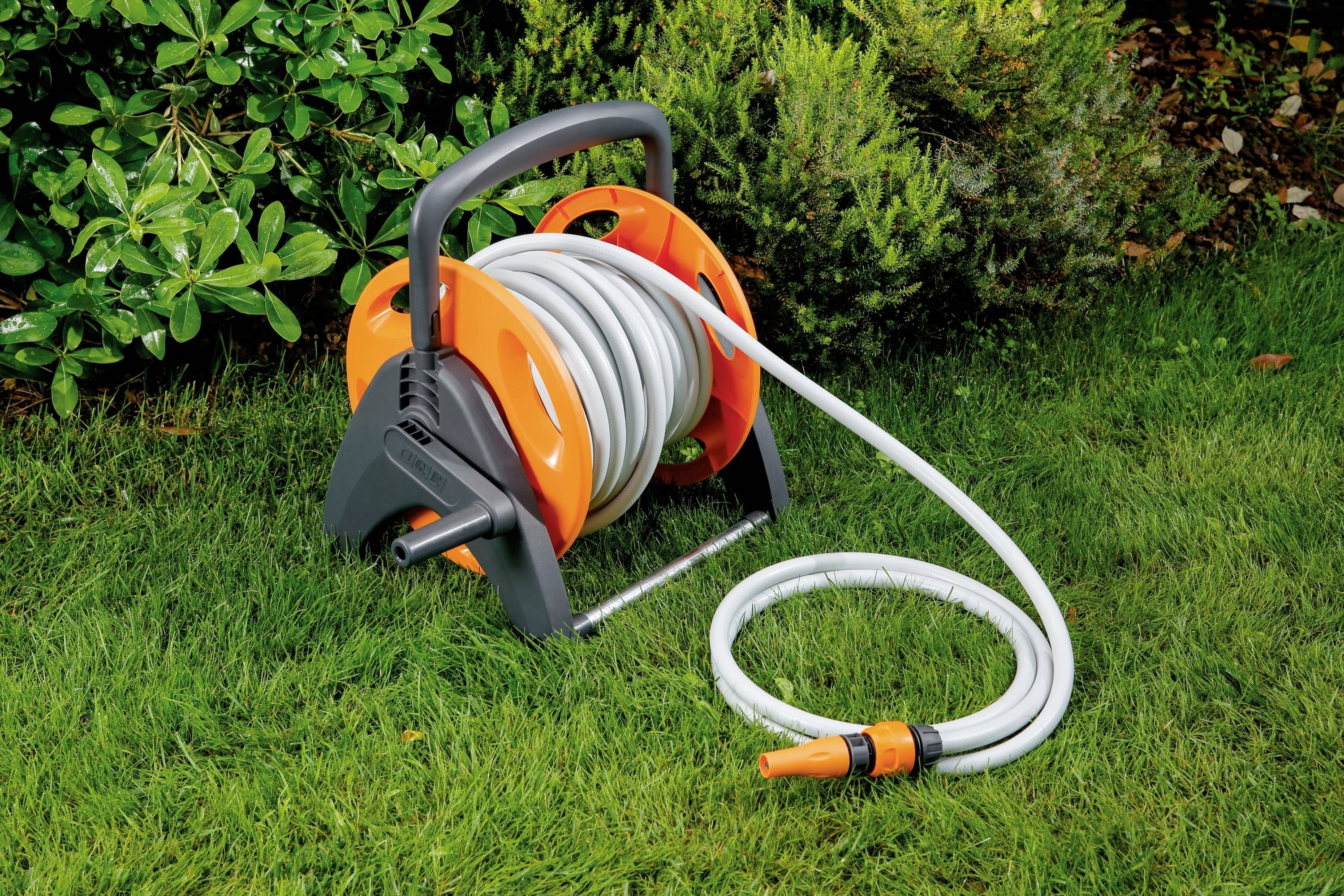 Een oranje-grijze tuinslang op een haspel ligt op groen gras, omgeven door struiken.