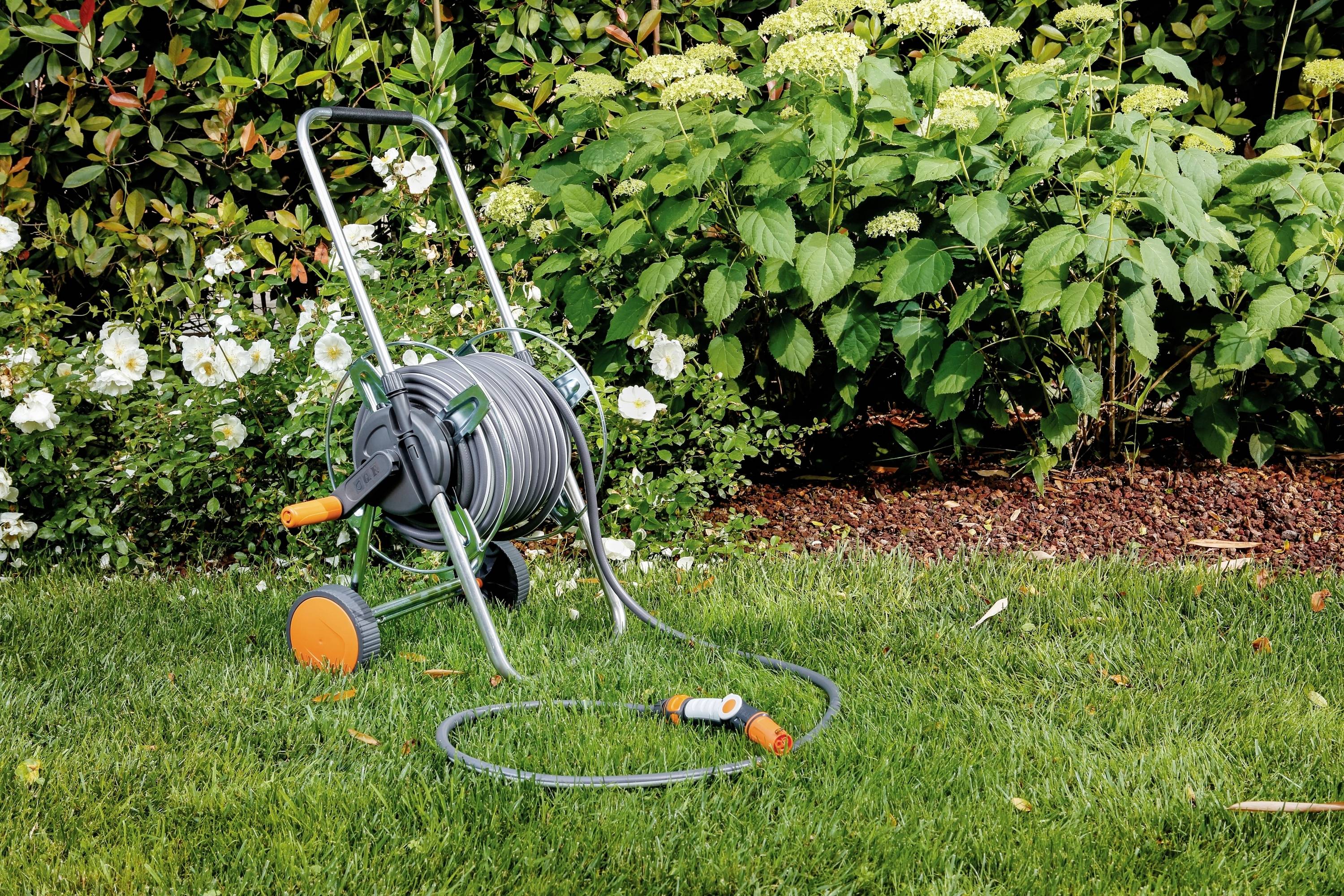 Een tuinslang op een slanghaspel staat op een goed onderhouden gazon. Op de achtergrond bevinden zich groene planten en witte bloemen.