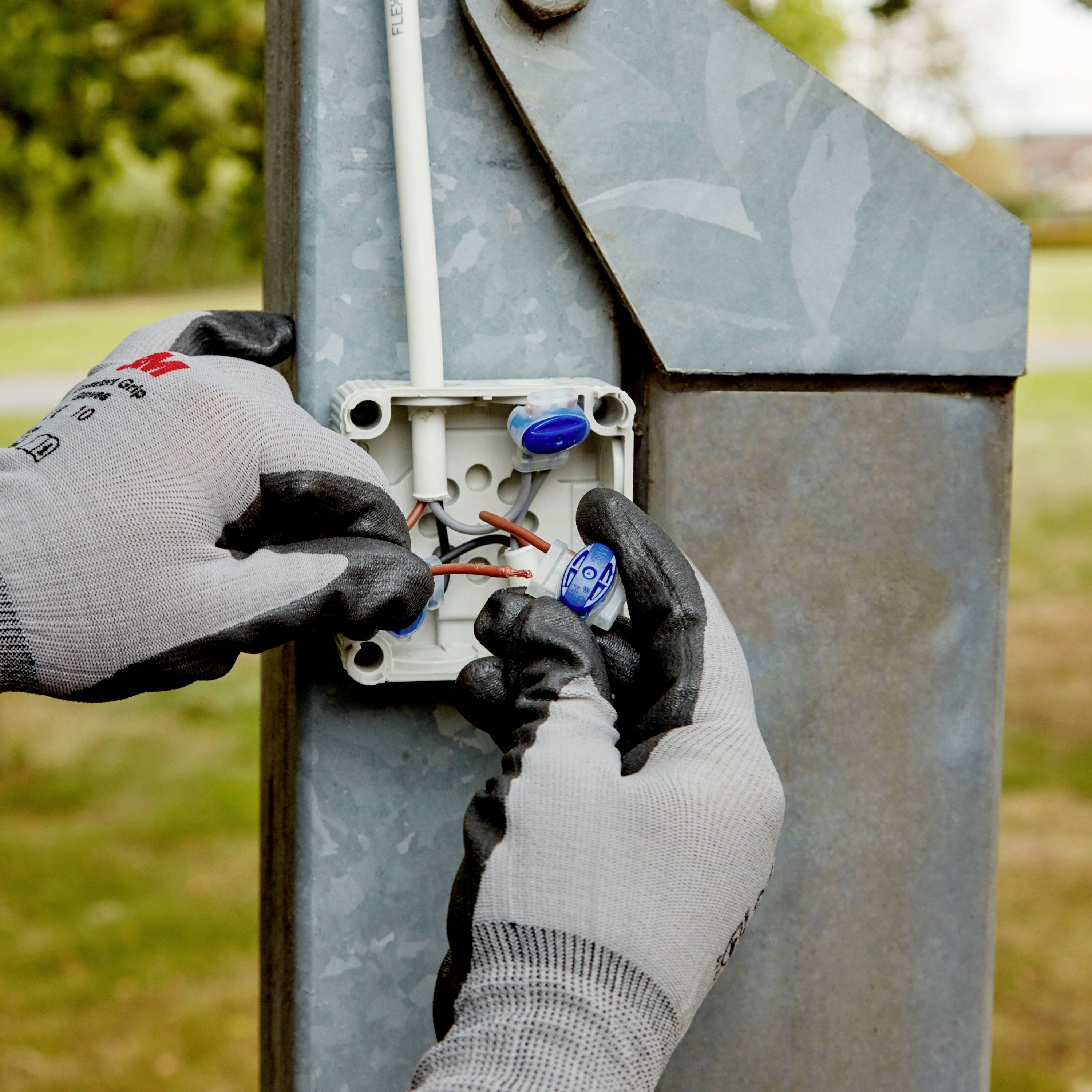 Een persoon met grijze handschoenen installeert een elektrische kabel in een elektriciteitskast buiten. Blauwe stekkerverbindingen zijn zichtbaar.