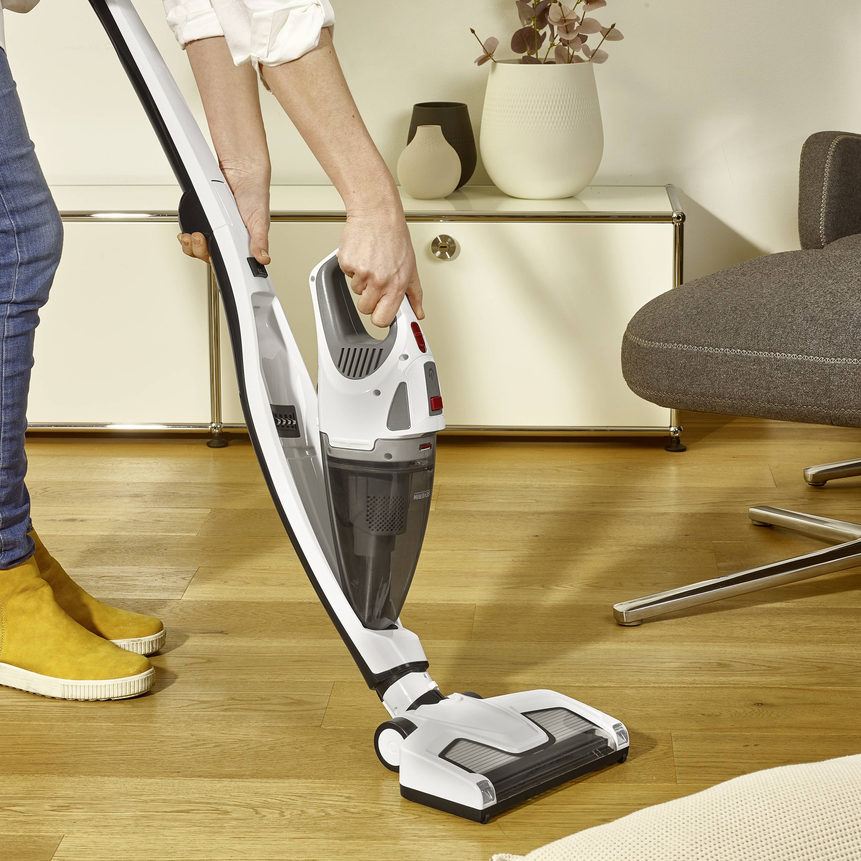 Een persoon zuigt met een draadloze stofzuiger een houten vloer in een gezellige ruimte; op de achtergrond staat een dressoir.