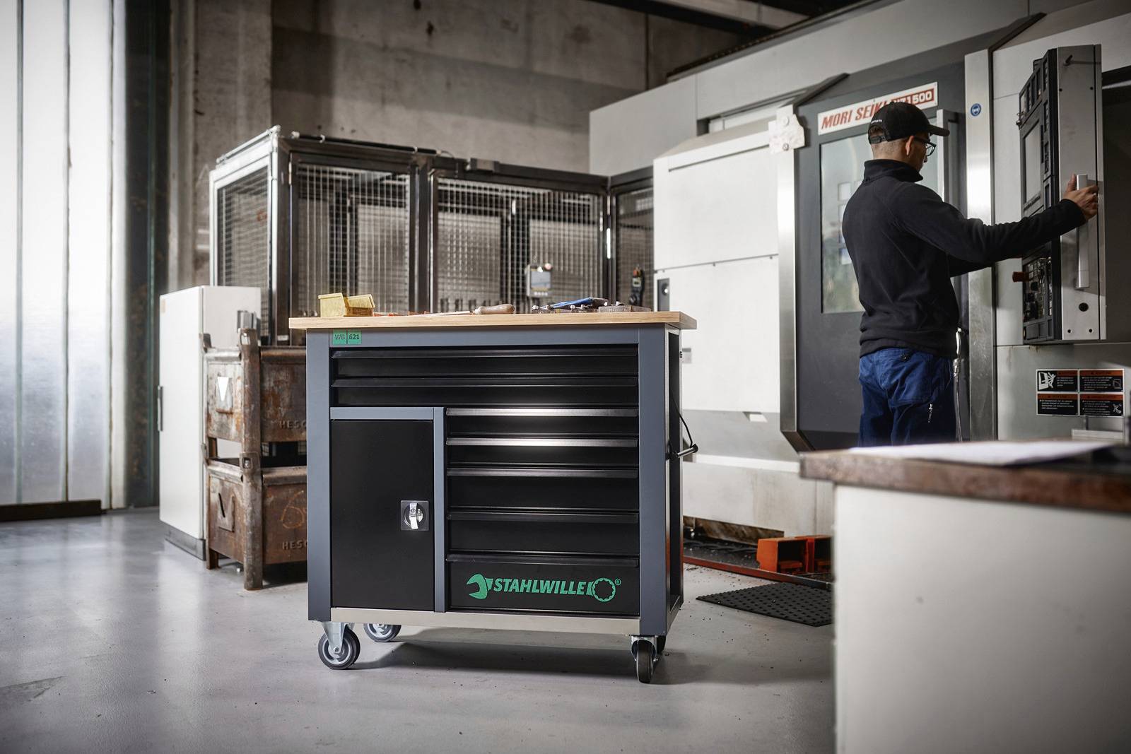Gereedschapswagen met merknaam staat in een werkplaats. Op de achtergrond bedient iemand een machine. Industriële omgeving.