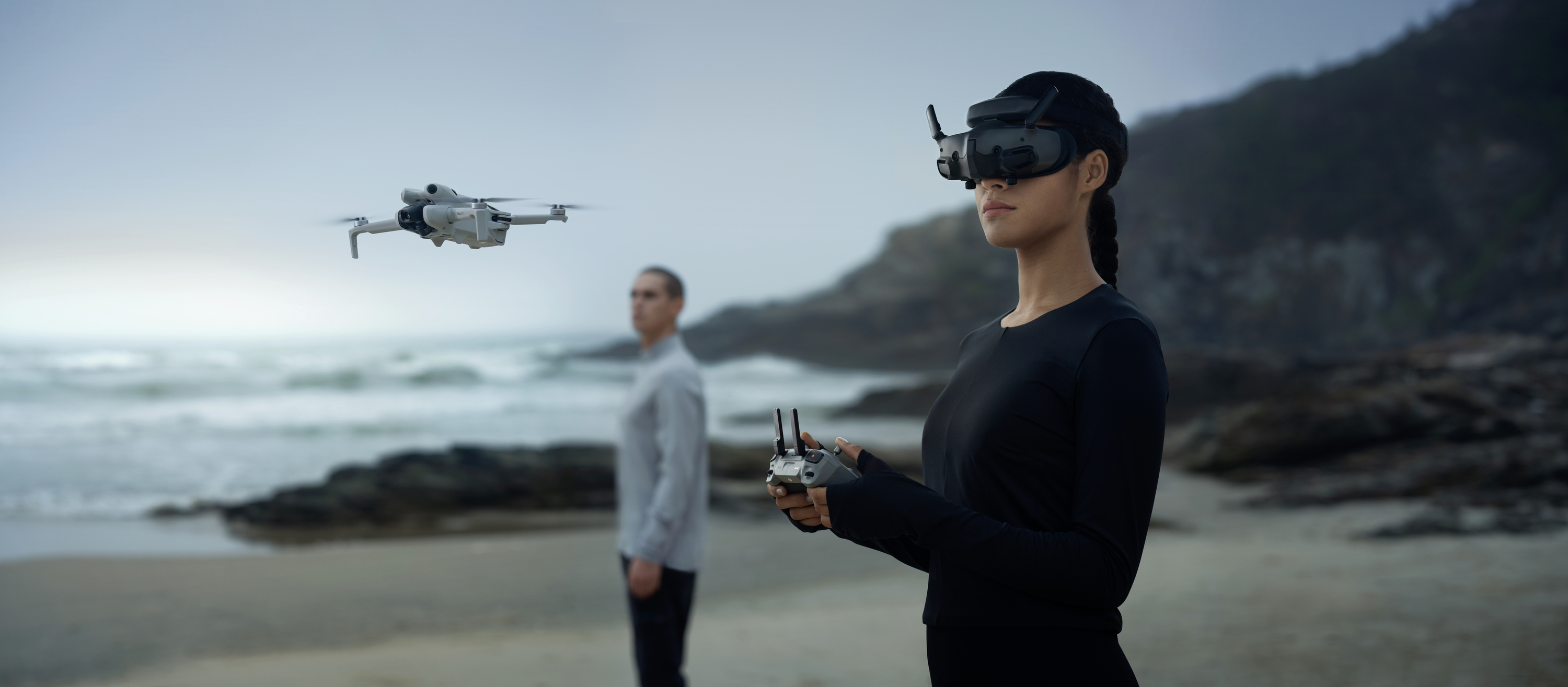 Een vrouw bestuurt een drone met een virtual-reality-bril op een strand, terwijl een man op de achtergrond staat.