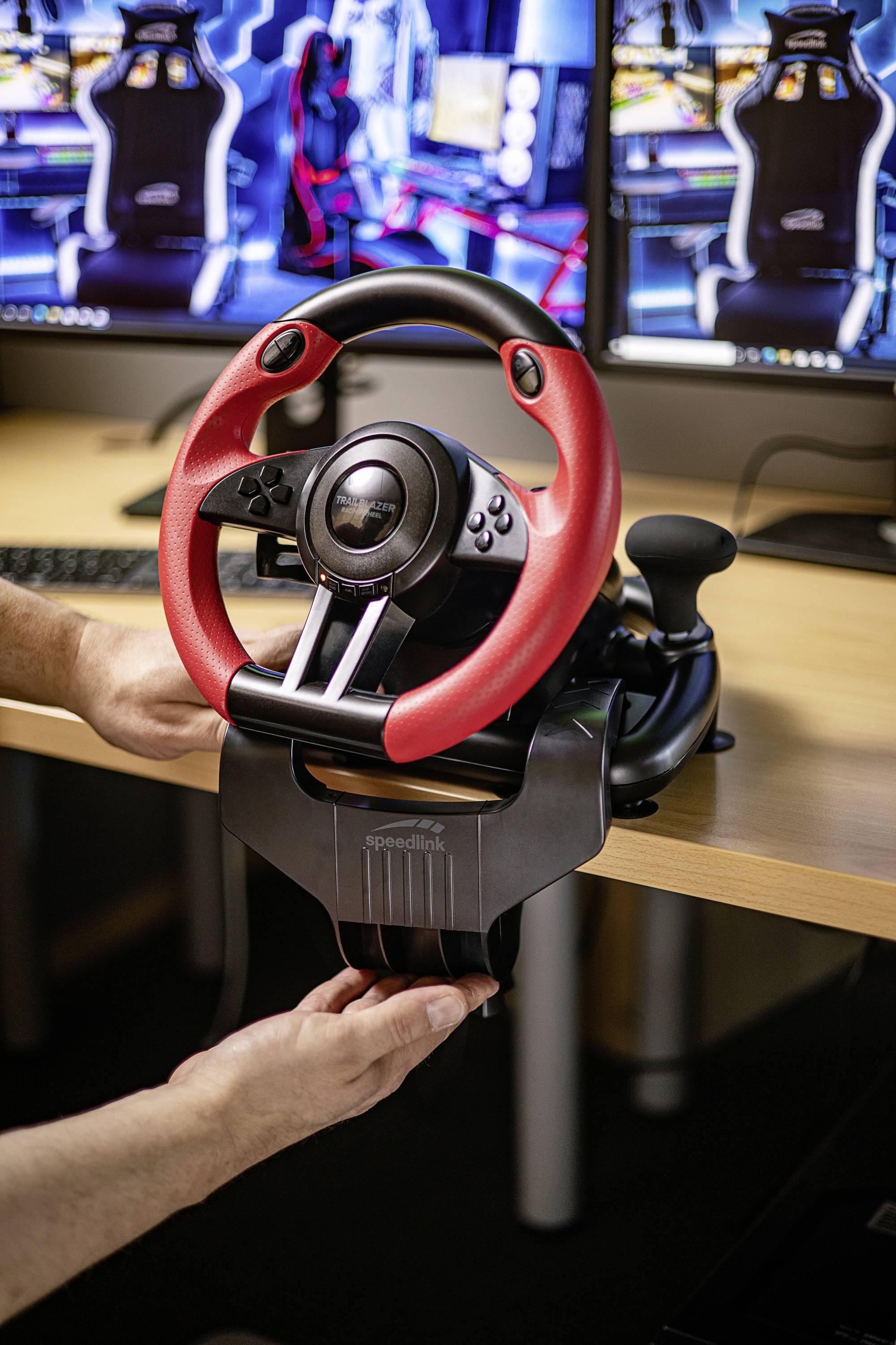 Een persoon monteert een rood en zwart racestuur aan een bureau, met twee monitors en gamingstoelen op de achtergrond.