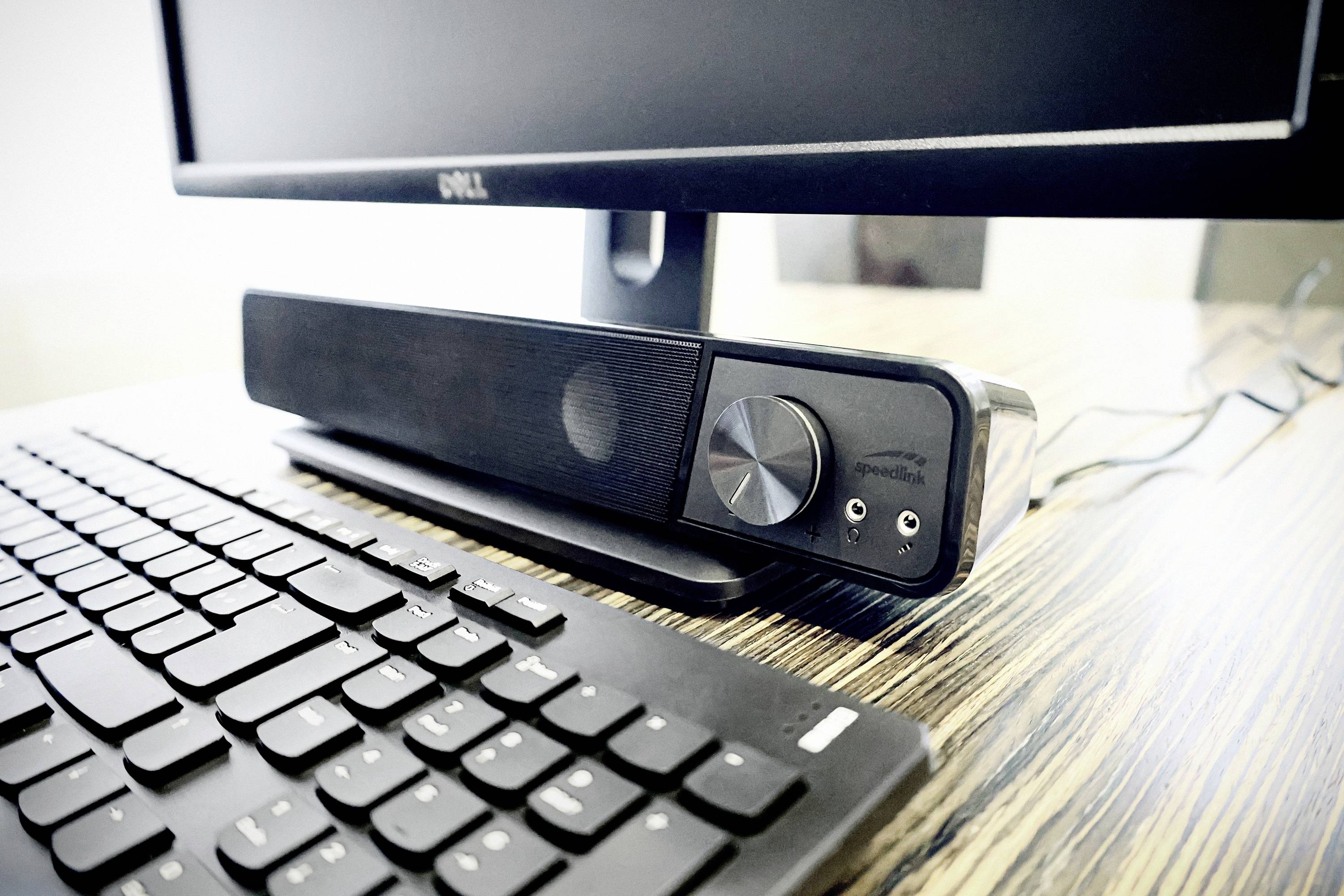 Een computerarbeidsplaats met een toetsenbord, een monitor en een soundbar op een houten tafel.