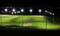 's Nachts verlicht voetbalveld met doelen, omringd door lichtmasten. Op de achtergrond zijn stadslichten zichtbaar.