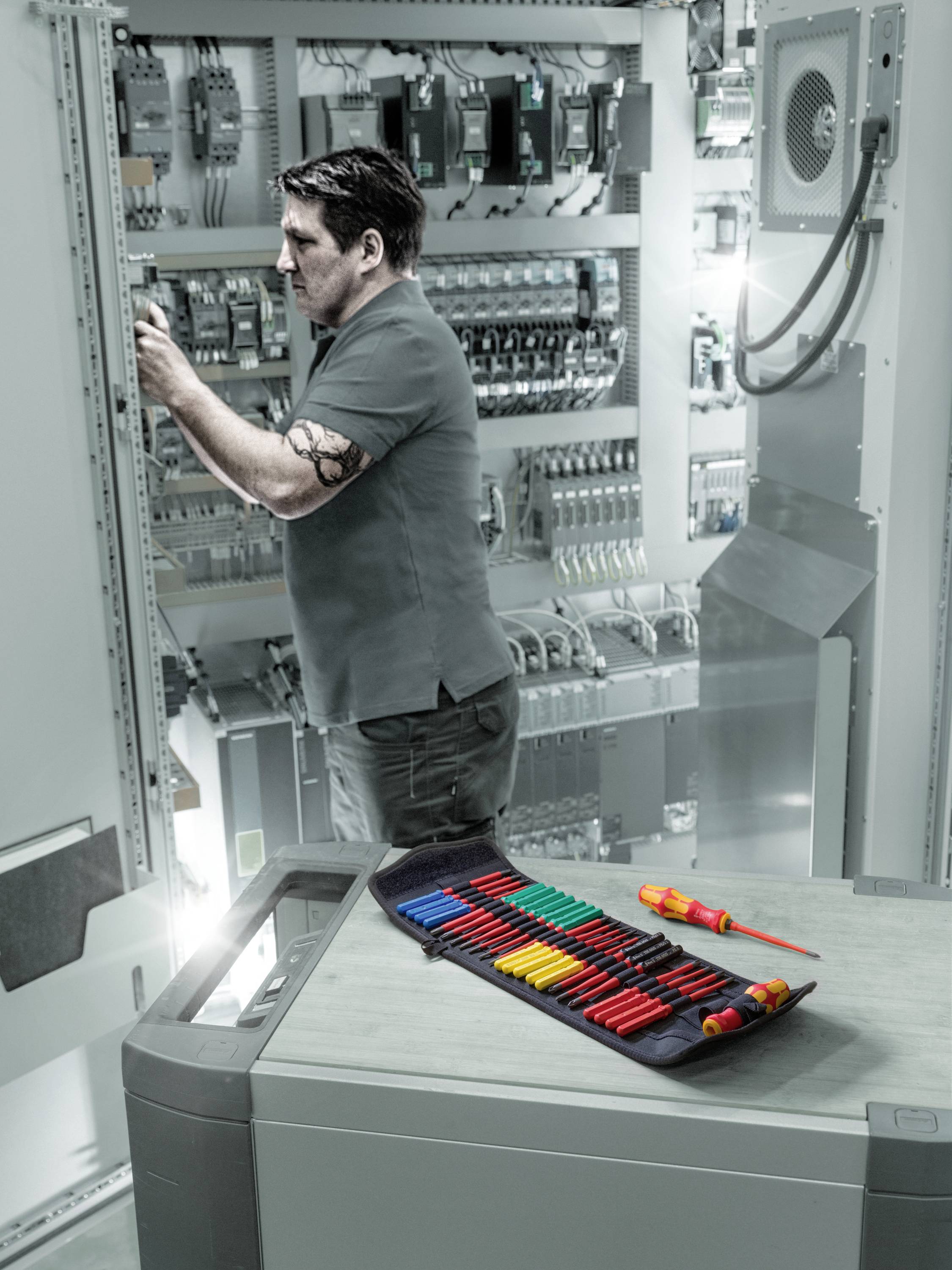 Een technicus werkt aan een schakelkast in een industriële omgeving. Op de voorgrond ligt een geopende etui met gereedschappen.
