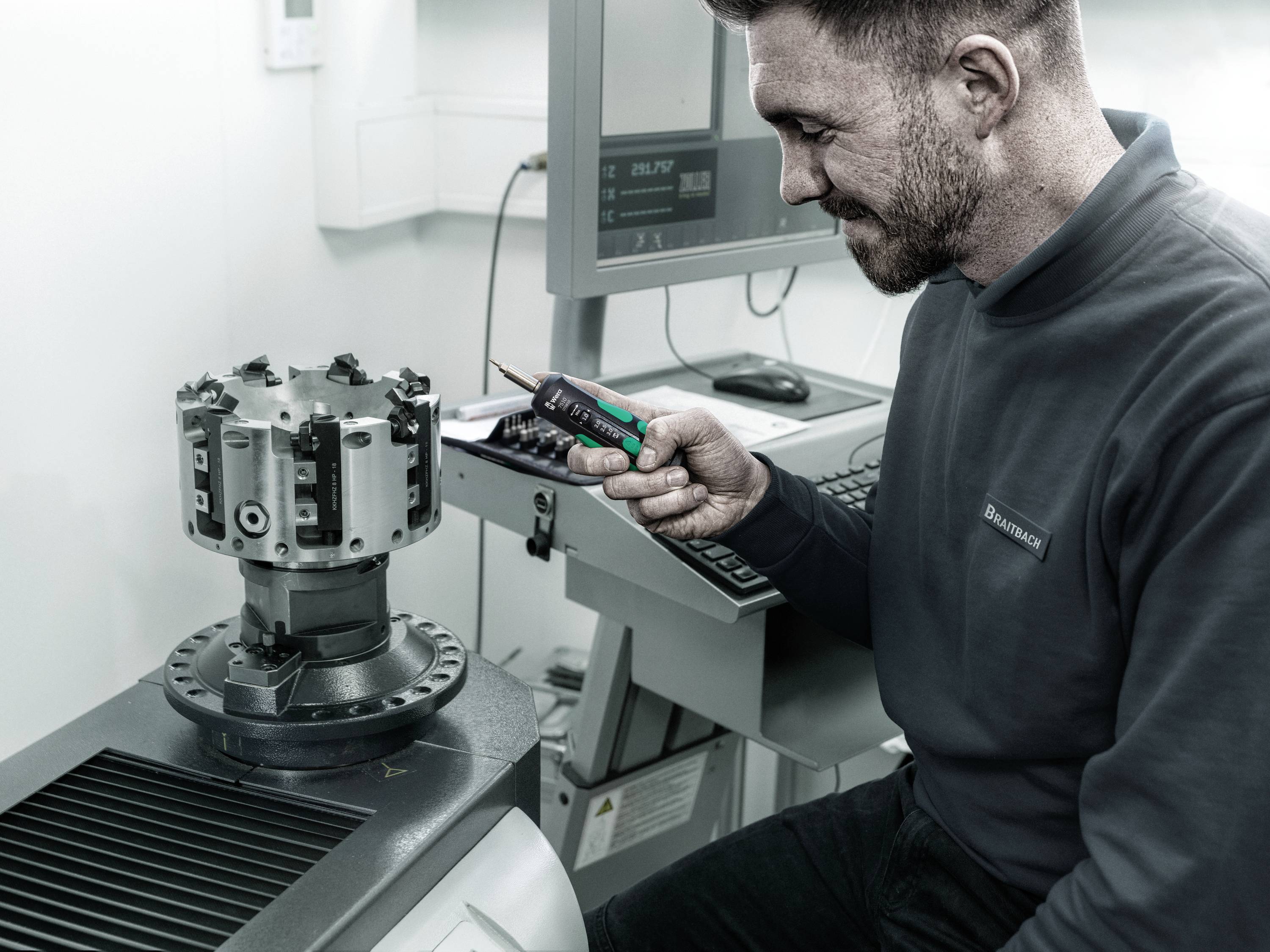 Een man meet een machinedeel met een digitaal meetinstrument in een laboratorium of werkplaats. Op de achtergrond staat een computer.