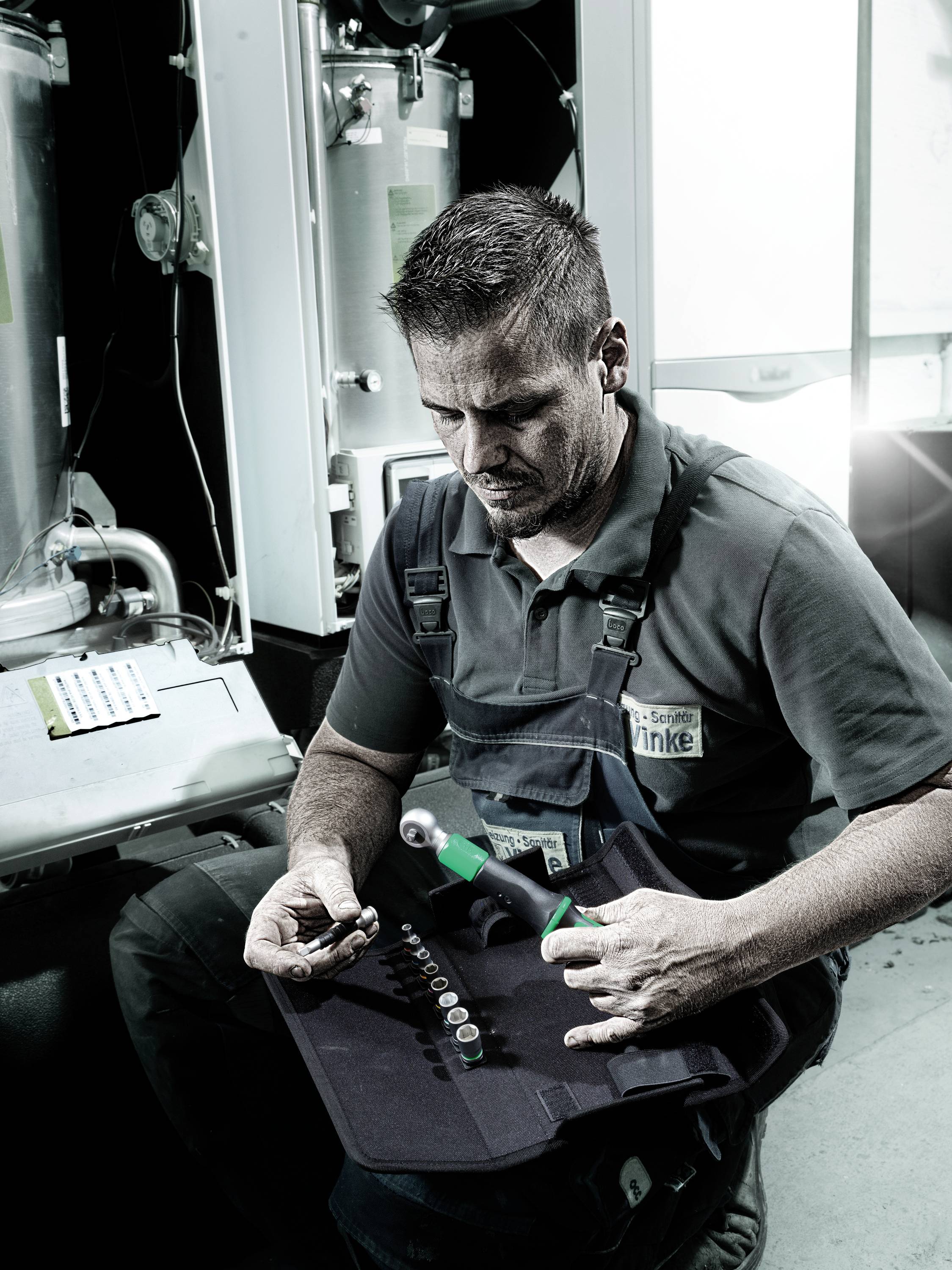 Een ambachtsman zit in een werkruimte en houdt gereedschap vast. Op de achtergrond zijn industriële machines zichtbaar.