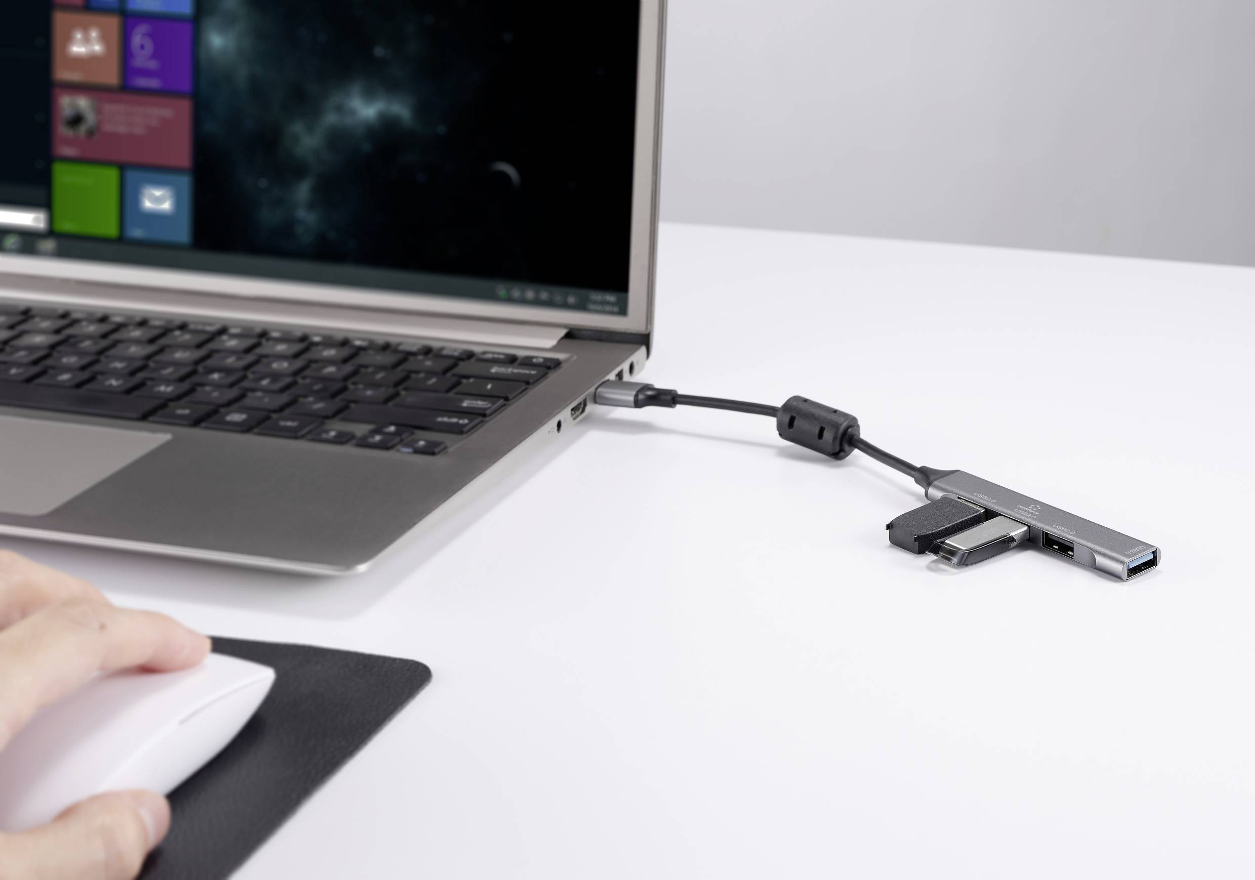 Een laptop met een open scherm staat op een tafel. Een USB-hub is aangesloten op de laptop. Een hand bedient een computermuis.