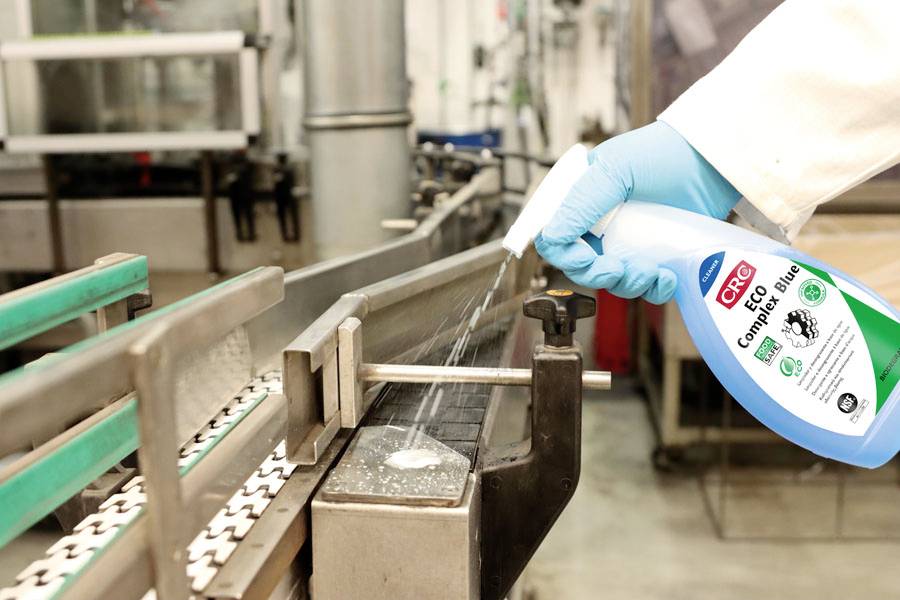 Een persoon met blauwe handschoenen sproeit een reinigingsmiddel op een transportband in een industriële omgeving.