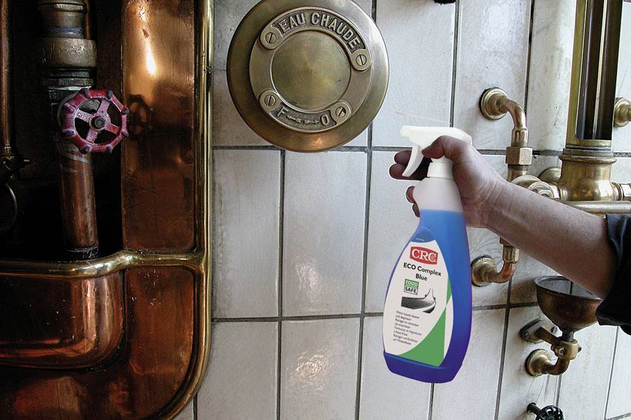 Een hand houdt een spuitfles voor oude leidingen en een waterkraan met de aanduiding 'Eau Chaude' (Heet water) aan de muur.