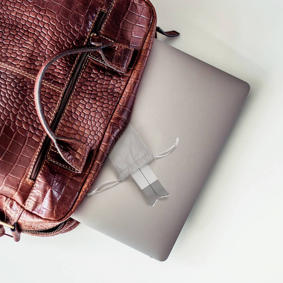 Een bruine krokodillenleren tas ligt op een zilveren laptop, die gedeeltelijk uit de tas steekt.