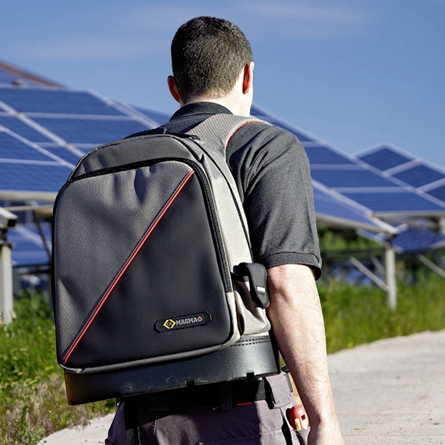 Een persoon met een zwart shirt draagt een grijze rugzak en staat voor zonnepanelen in de buitenlucht.