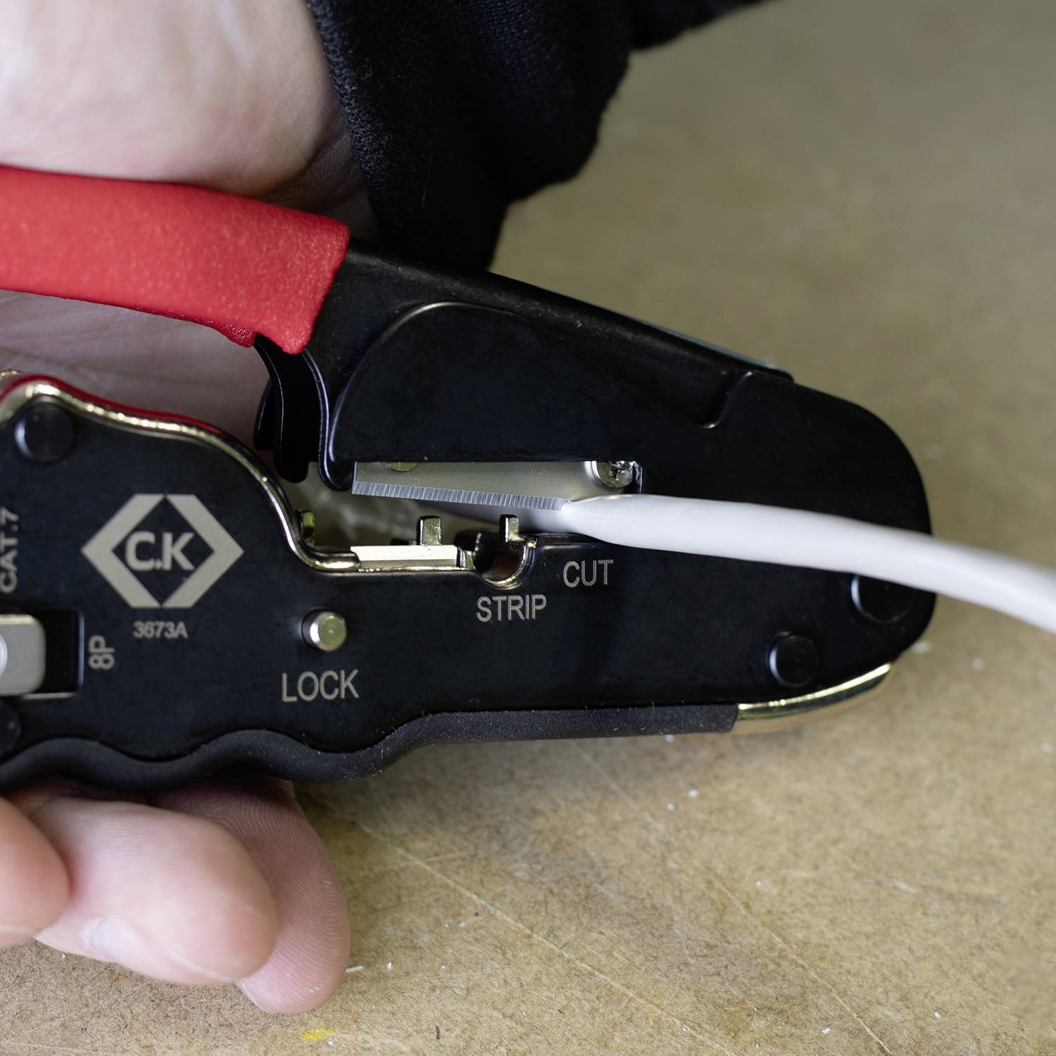 Een hand houdt een kabelstripper vast die een wit kabel doorsnijdt. De kabelstripper toont de markeringen 'Strip' en 'Cut'.