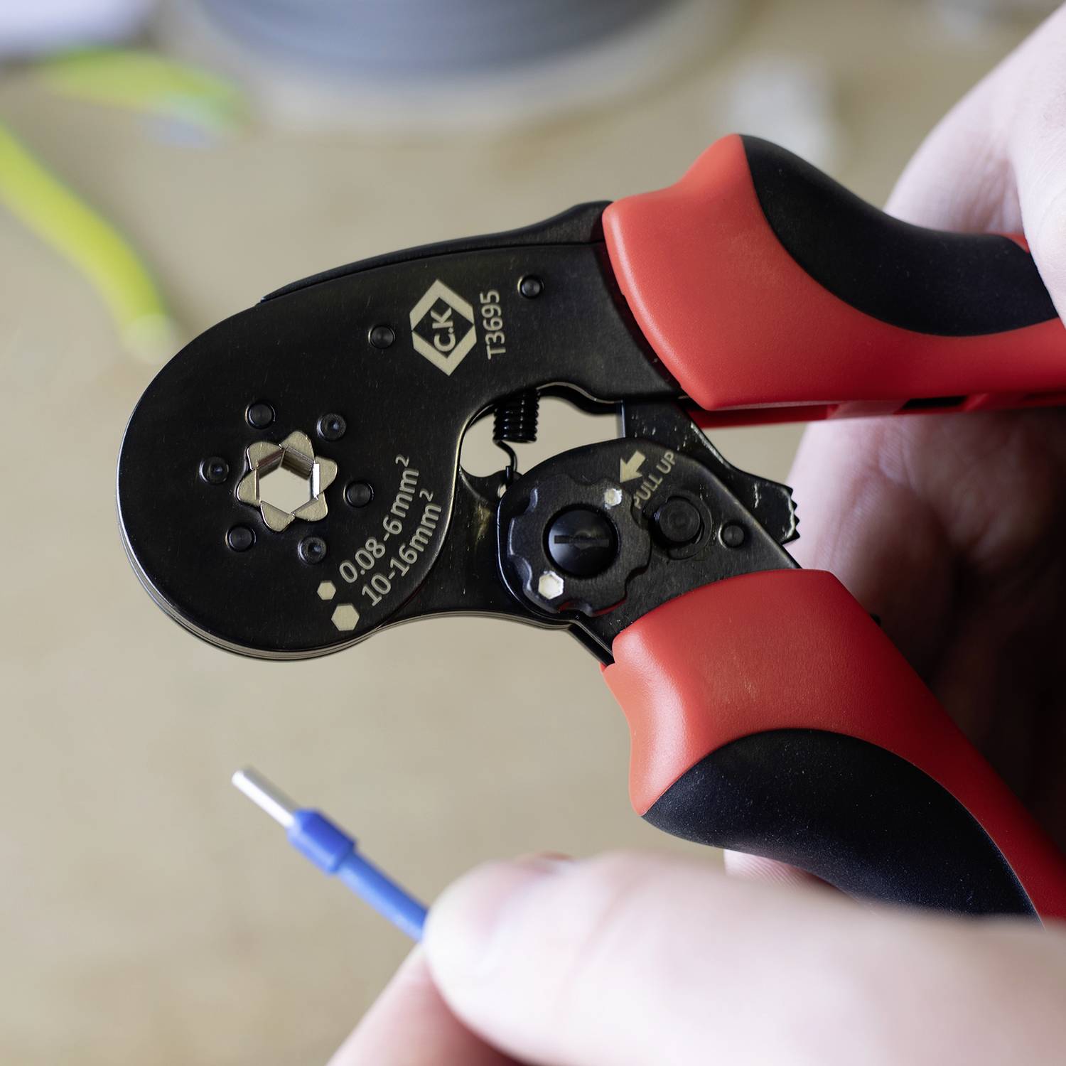 Een hand houdt een rode krimptang vast, die werkt aan een blauw kabel.