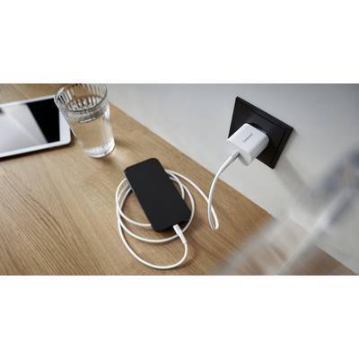 Een smartphone ligt op een houten tafel en wordt opgeladen via een wit snoer dat in het stopcontact steekt. Ernaast staat een glas water en een tablet.