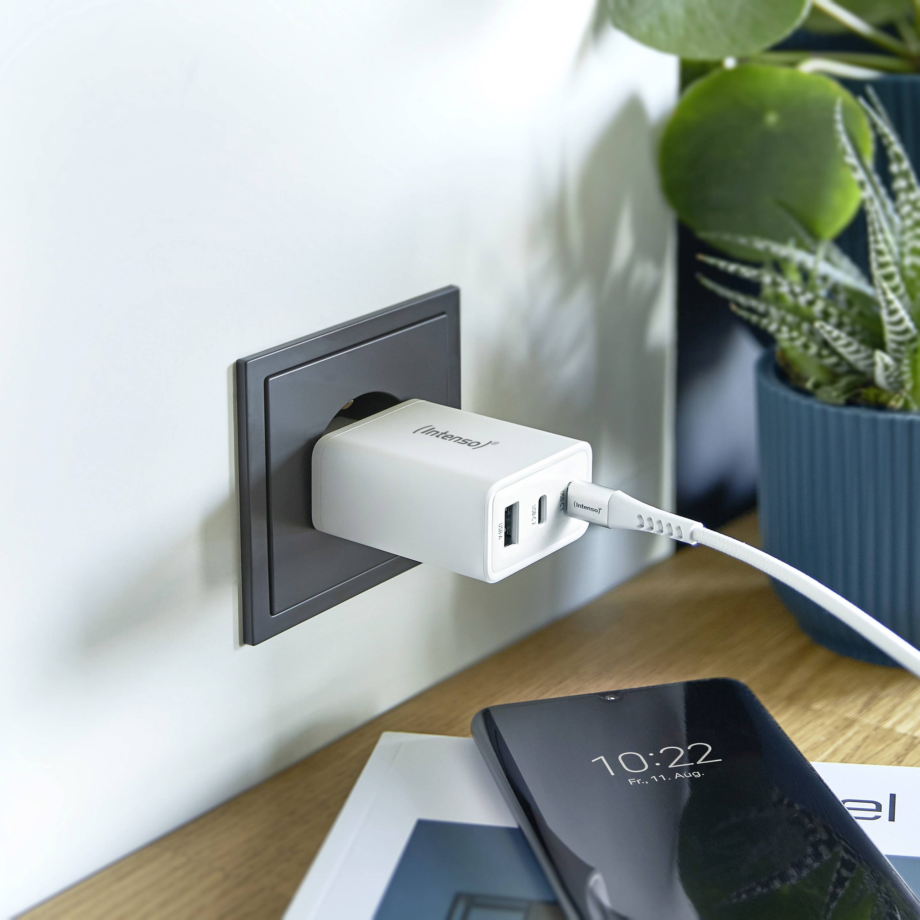 Een smartphone wordt via een oplader opgeladen aan een stopcontact. Ernaast staat een bloempot.