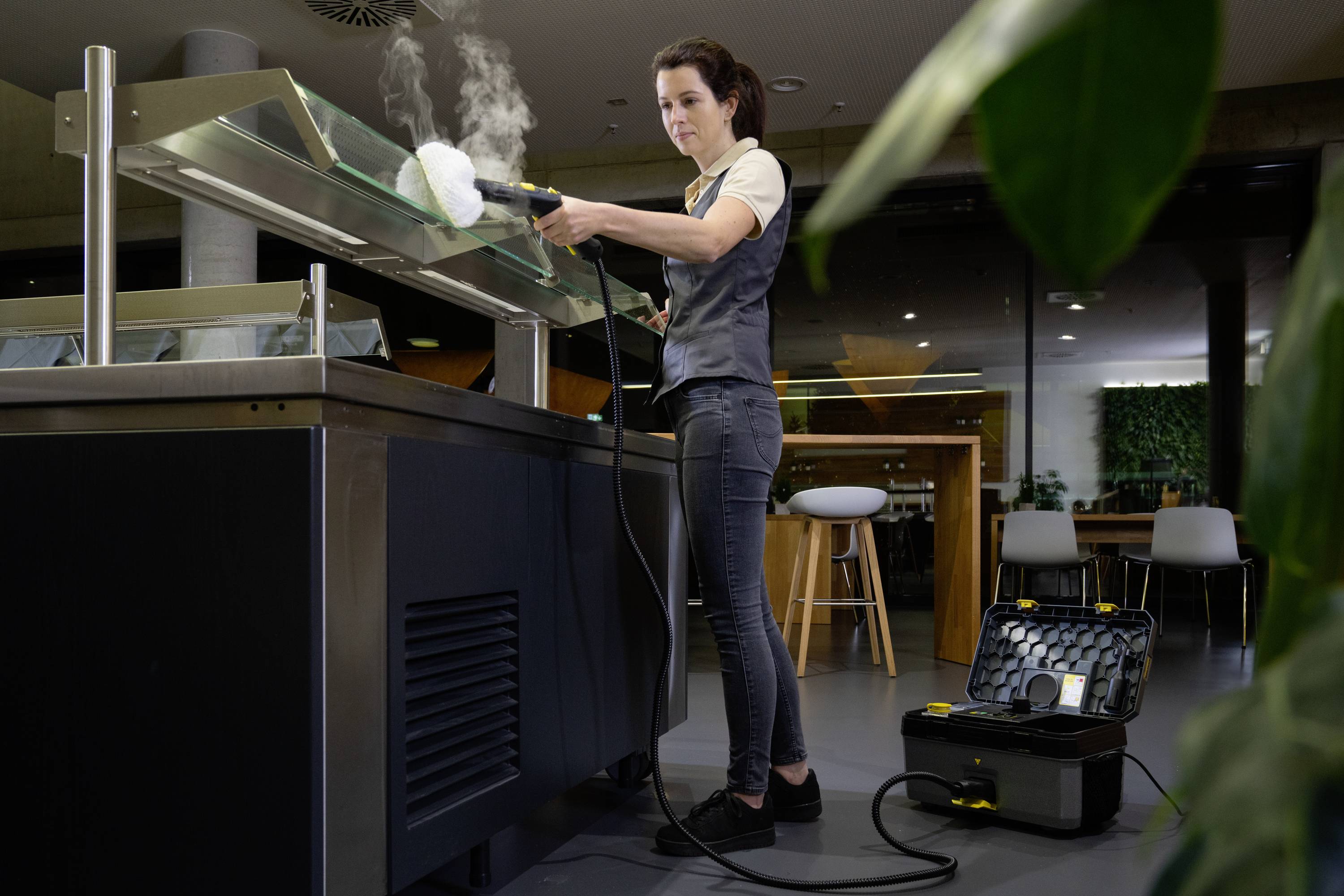 'Een vrouw reinigt het glazen oppervlak van een buffetkast met een stoomreiniger in een moderne eetzaal. De reinigingsapparatuur en toebehoren zijn zichtbaar.'