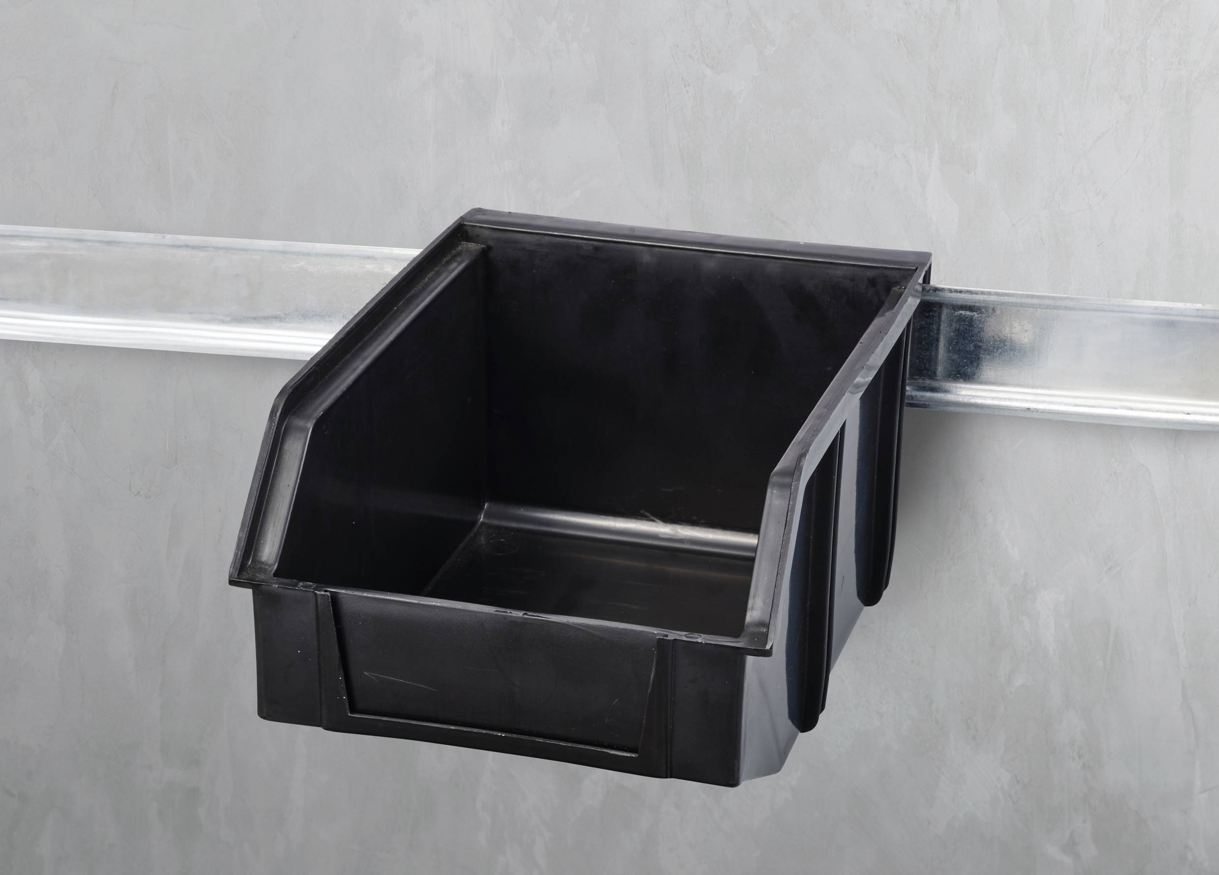 Zwarte kunststof opbergdoos, opgehangen aan een metalen rail aan de muur. De doos is leeg.