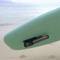 Een groen surfboard staat rechtop op het strand met de zee op de achtergrond. Aan het board is een zwart voorwerp bevestigd.