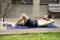 Een man ligt op een yogamat en doet sit-ups in de buitenlucht, met een glimlach op zijn gezicht. Op de achtergrond is een bank te zien.