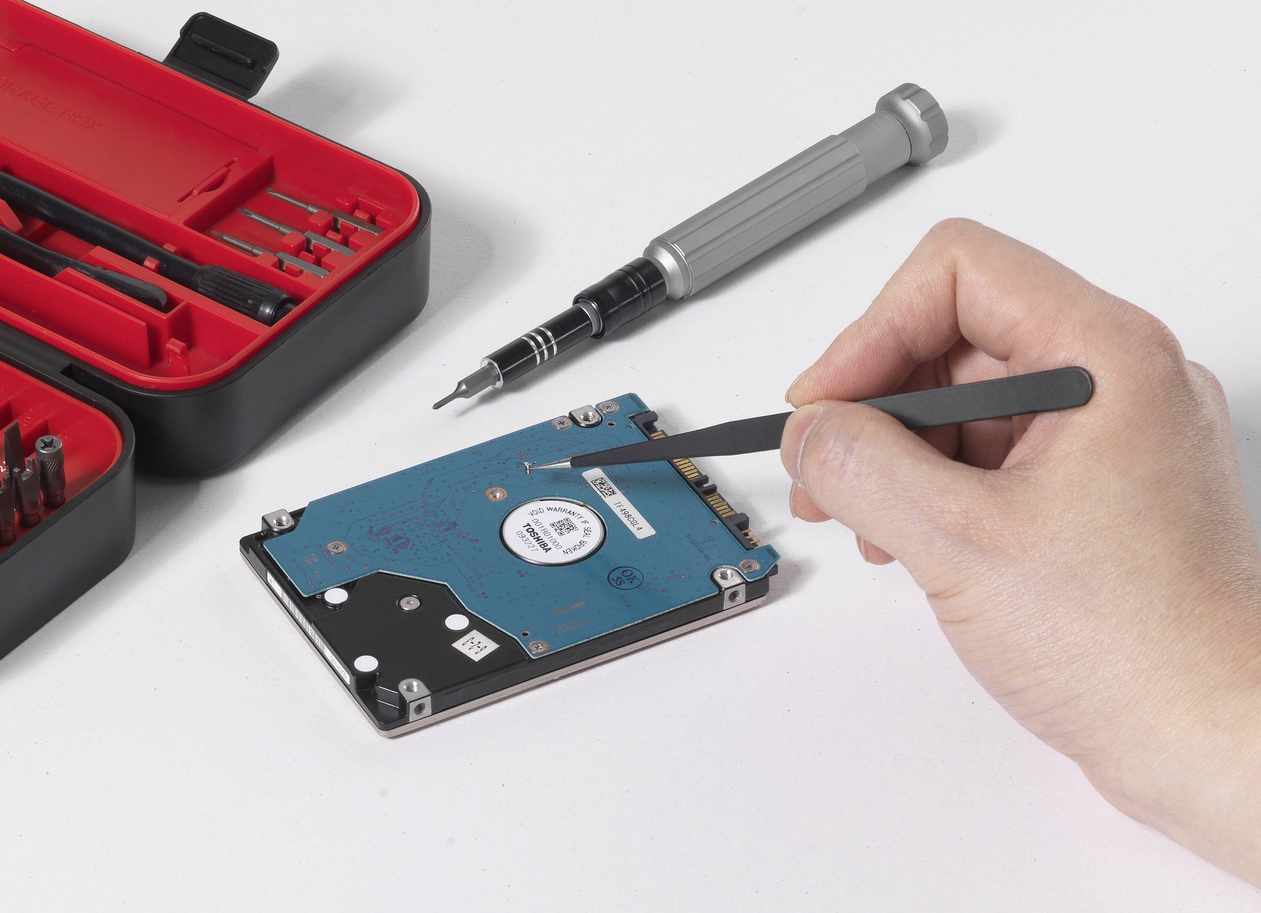 Een hand gebruikt een pincet om een computerharddisk te repareren. Op de achtergrond bevindt zich een openliggende schroevendreier-set.